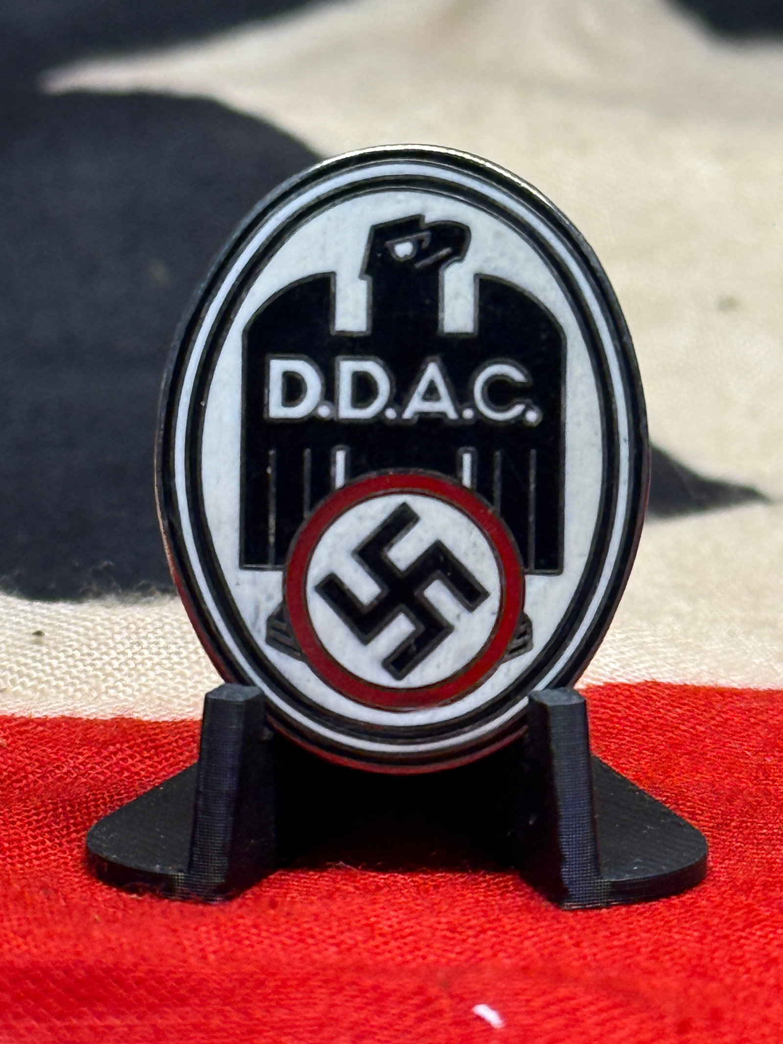 WWII German D.D.A.C. Eagle & Swastika Enamel Membership Pin: An oval WWII German enamel pin displaying a bold black eagle motif against a white field, with the initials “D.D.A.C.” prominently positioned across the center and a black swastika enclosed wi