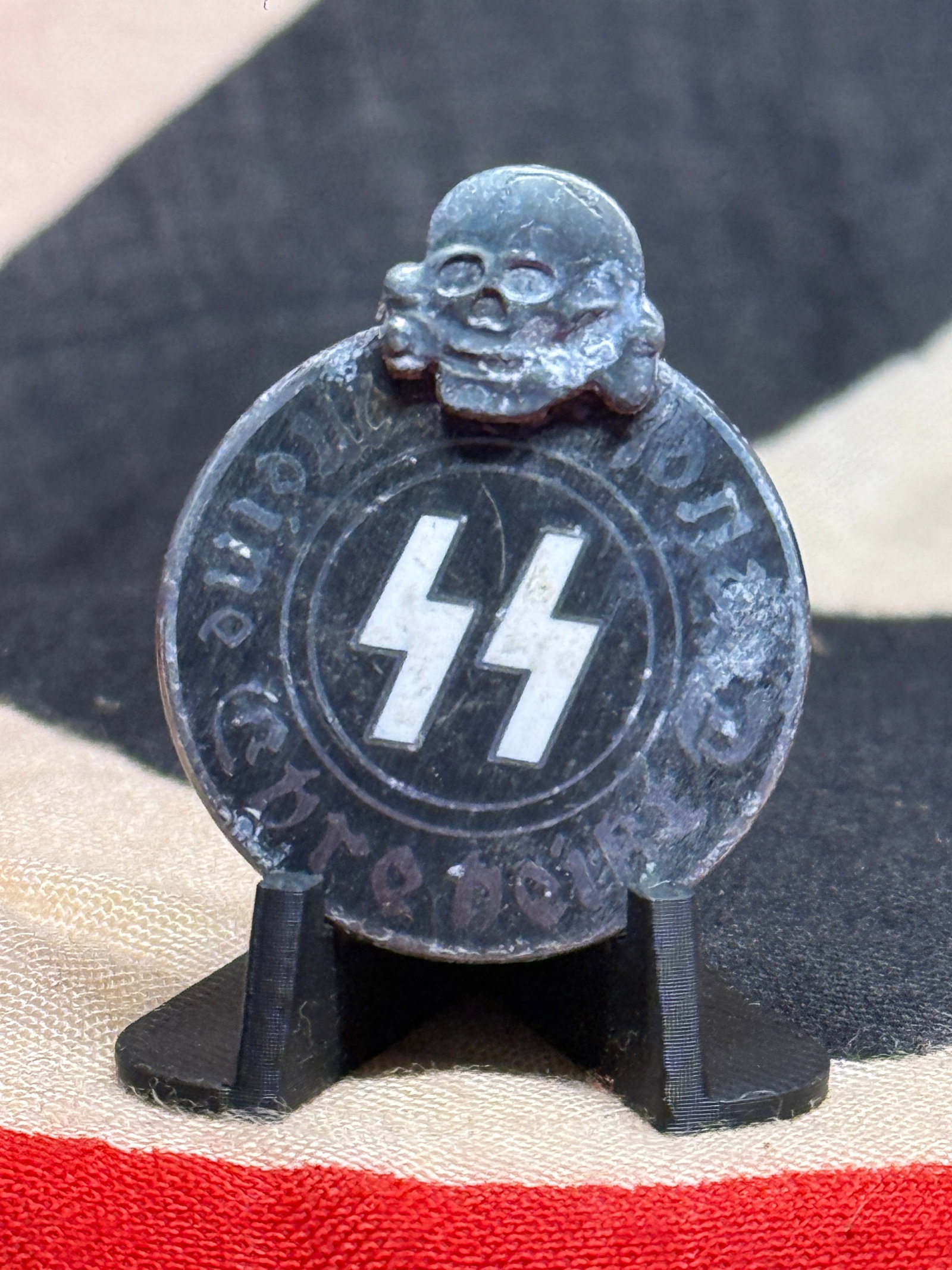 WWII German SS Totenkopf & SS Runes Membership Pin – RZM Marked: This unusual WWII German SS-associated pin features a circular dark-colored face centered by prominent white SS runes and surmounted by a raised Totenkopf skull-and-crossbones device. Faint German let