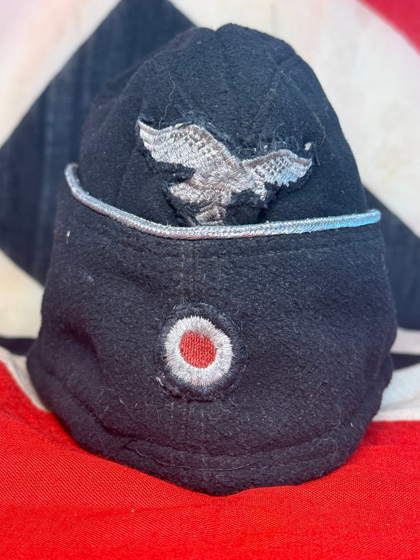 WWII German Luftwaffe Black Overseas Side Cap with Silver Piping, Eagle & Cockade – RZM Label: This distinctive WWII German black wool overseas side cap features silver-colored piping following the upper edge, together with an embroidered national eagle positioned prominently on the front and a