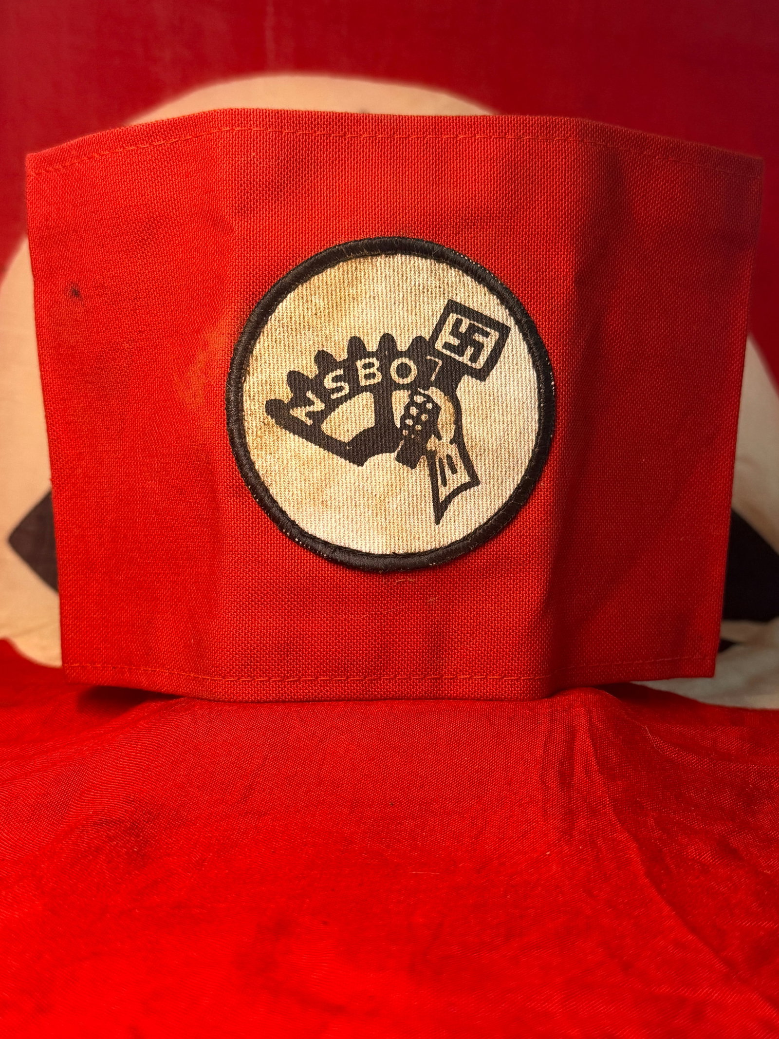 WWII-Era German NSBO National Socialist Factory Cell Organization Armband: This striking red textile armband bears a large circular white central patch outlined in black and printed with the emblem and initials “NSBO,” representing the Nationalsozialistische Betriebs