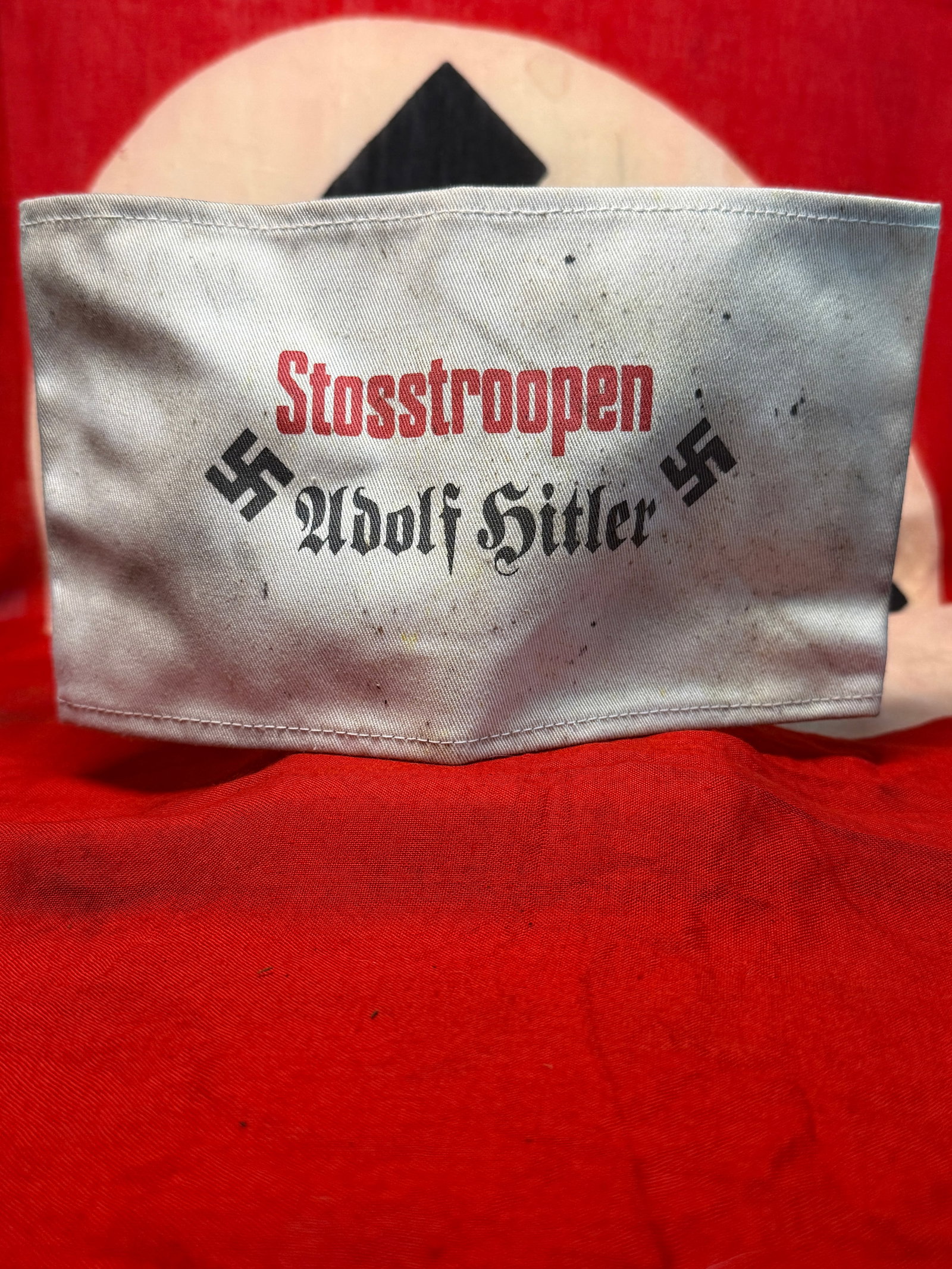WWII German “Stosstruppen Adolf Hitler?? Swastika Armband: A distinctive light-gray textile armband prominently printed with the red word “Stosstruppen” above the black Gothic inscription “Adolf Hitler,” flanked on either side by black swastik