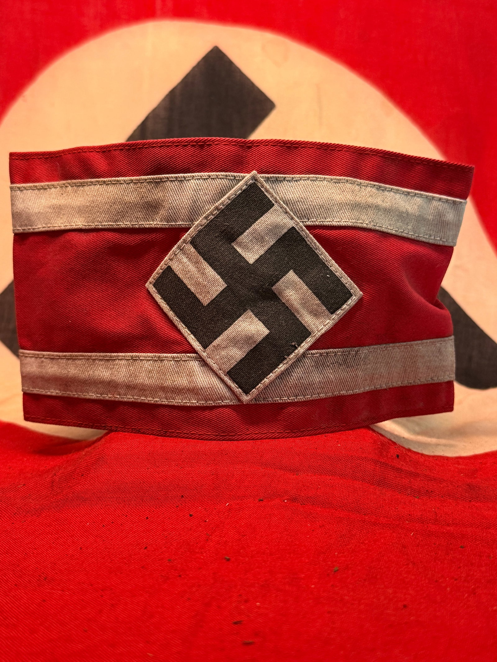 WWII-Era German Red Swastika Armband with Silver-Gray Stripes & RZM Label: A distinctive German organizational armband constructed from deep red fabric with two horizontal silver-gray bands and a centrally applied diamond-shaped panel bearing a large black swastika. The sepa