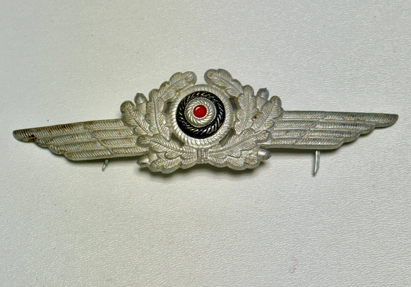 WWII German Luftwaffe Pilot’s Metal Winged Wreath & Cockade Insignia: WWII German Luftwaffe metal insignia featuring an impressive horizontally winged design centered by a raised oak-leaf wreath and national cockade in black, silver-tone, and red. The deeply detailed co
