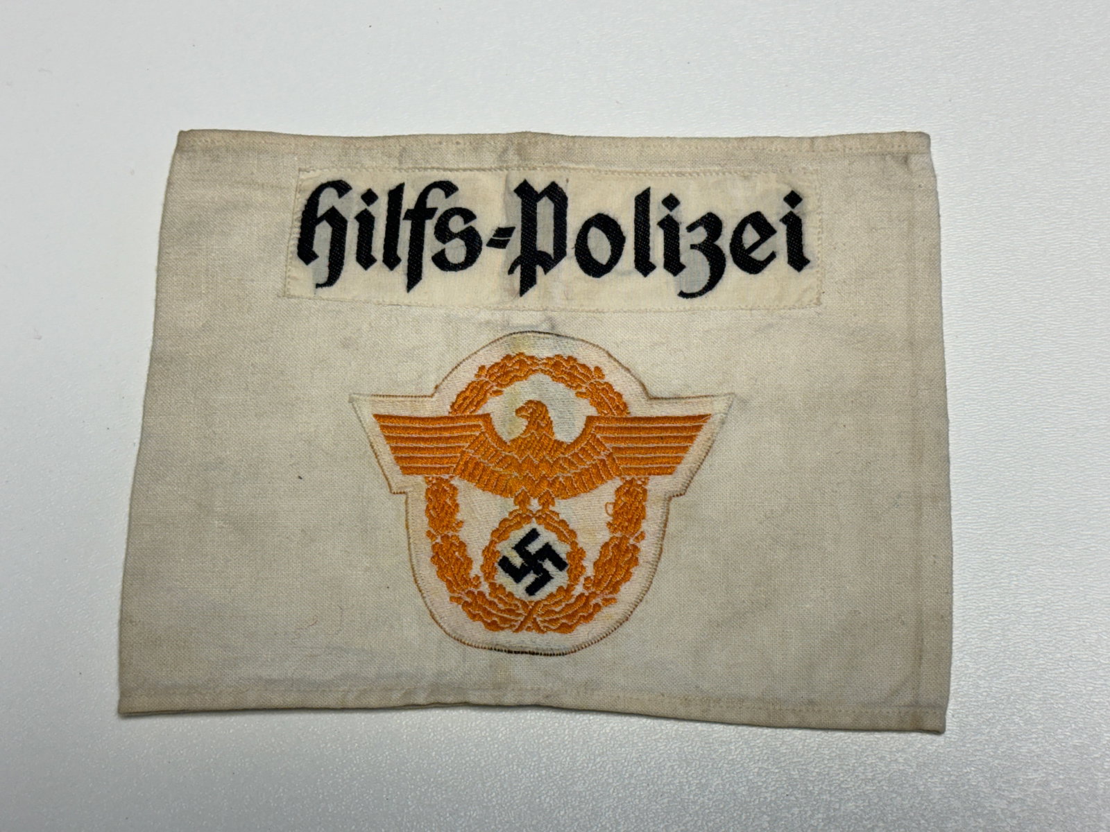 WWII German Hilfspolizei Auxiliary Police Embroidered Armband: WWII German Hilfspolizei auxiliary police cloth armband constructed of off-white fabric and featuring a prominent black Gothic-script “Hilfs-Polizei” designation across the upper portion. The 