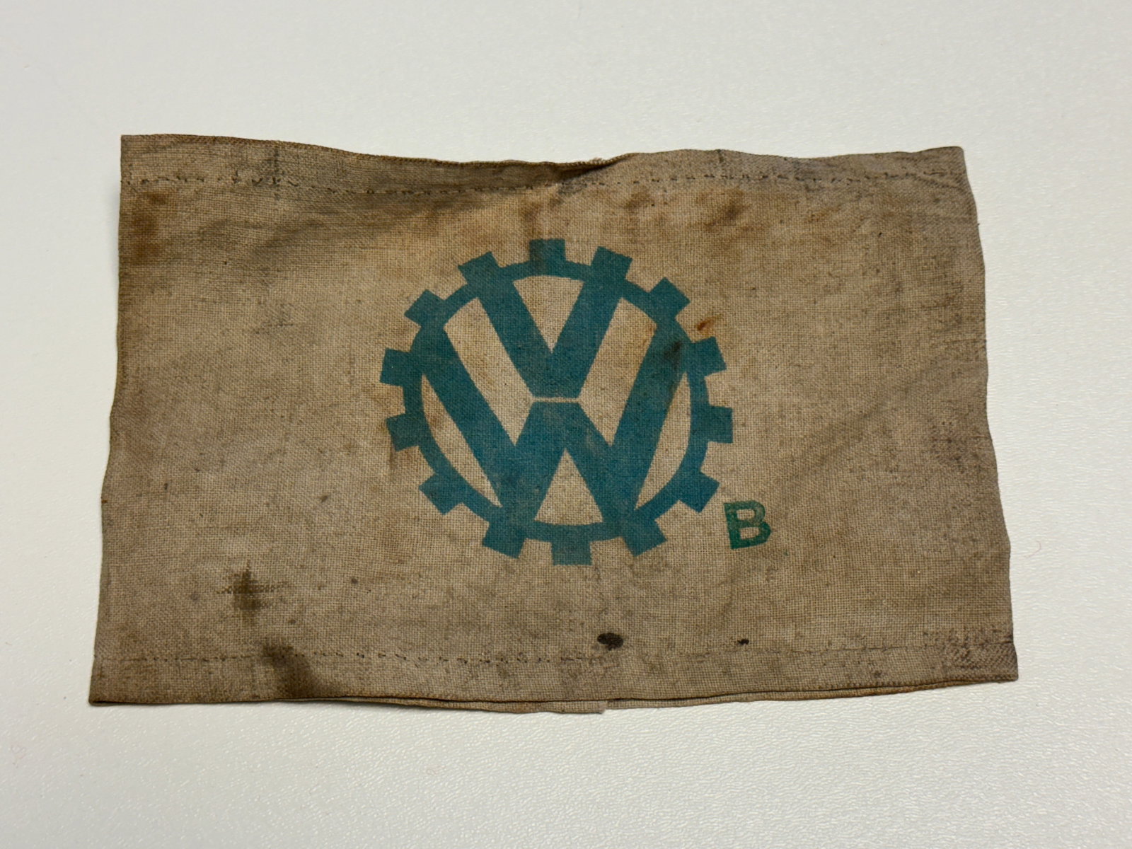 WWII German Volkswagen VW Factory Worker Cloth Armband – Wolfsburg Automotive Industry: WWII German cloth armband associated with Volkswagen industrial and factory personnel, constructed from coarse tan woven fabric and prominently printed with the distinctive blue-green “VW” mon