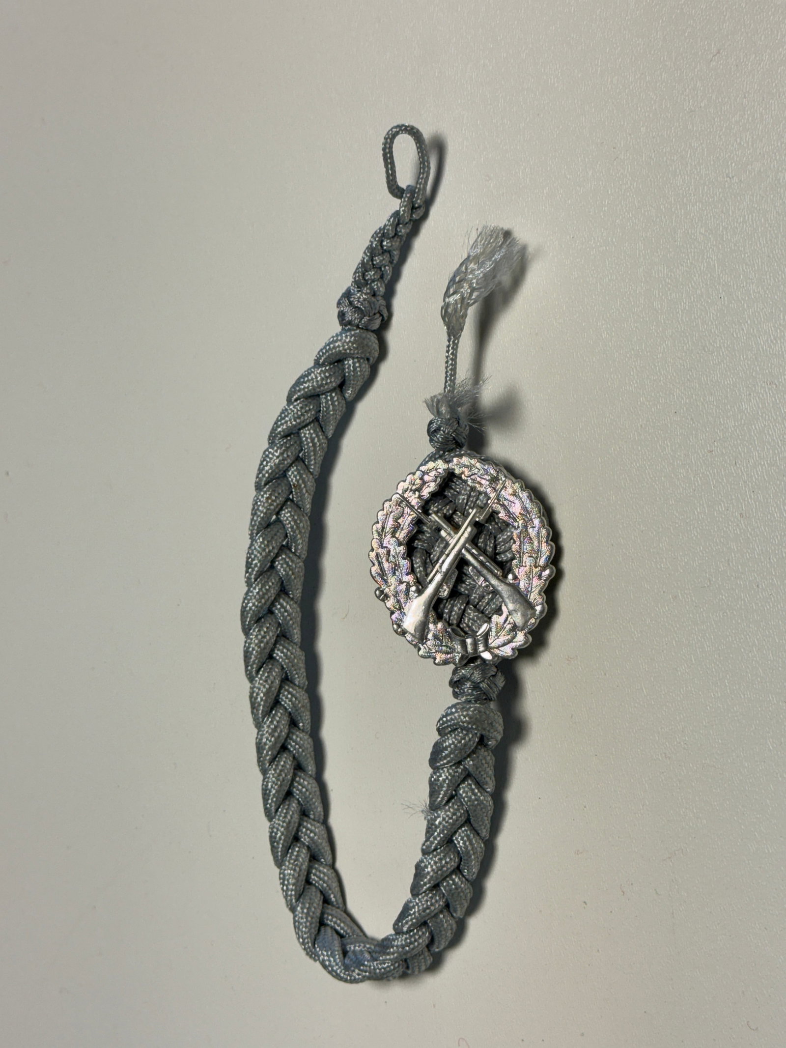 WWII German Army Infantry Marksmanship Lanyard with Rifle Badge: WWII German military marksmanship lanyard constructed of braided gray cord and fitted with an oval metal shooting badge featuring a detailed oak-leaf wreath surrounding crossed rifles, with a tied bow