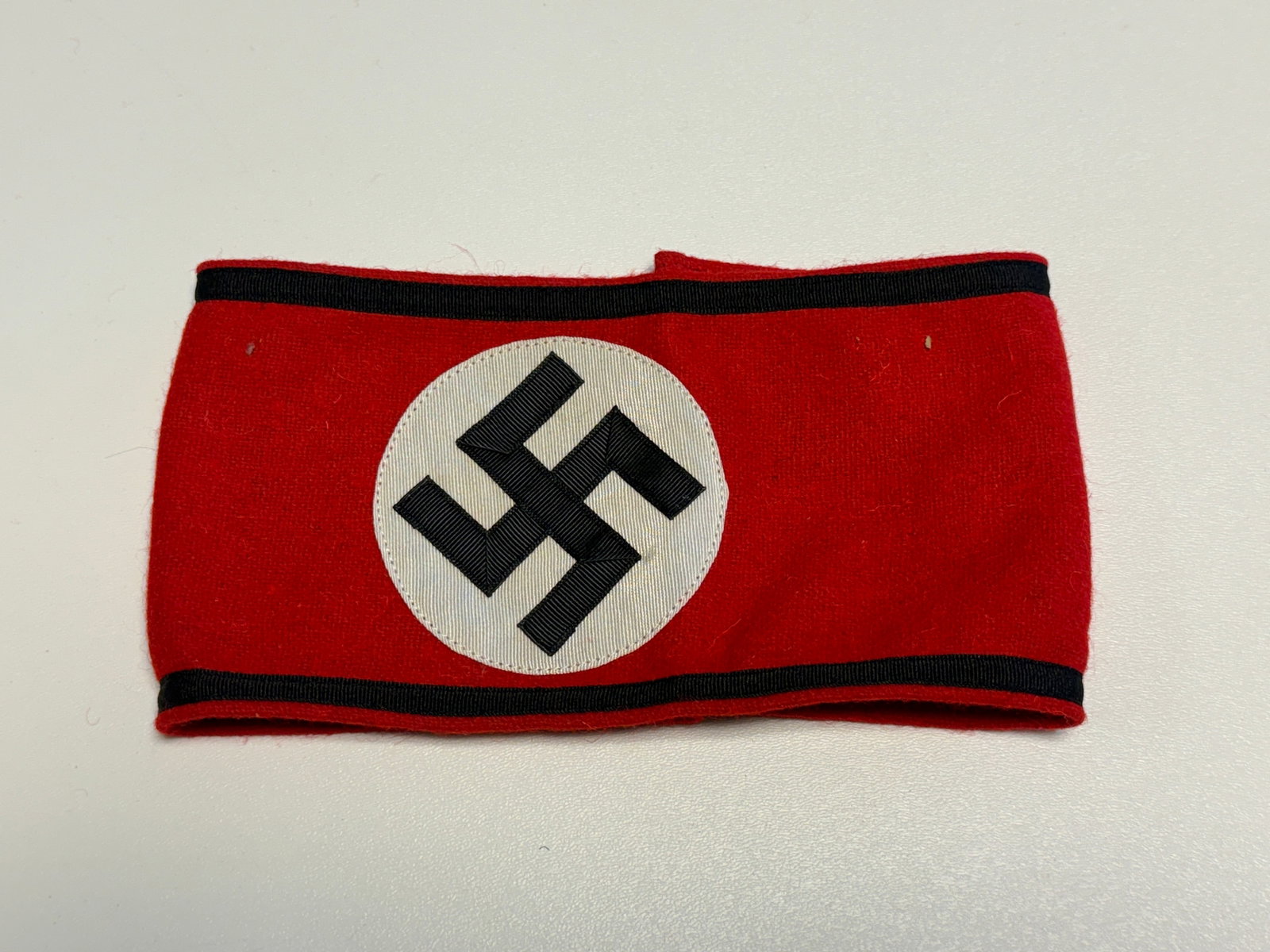 WWII German SS Officer RZM Wool Armband with Black Borders: WWII German SS officer armband constructed of red wool with black woven borders along the upper and lower edges and a central white circular field bearing a black swastika. The interior retains sewn b