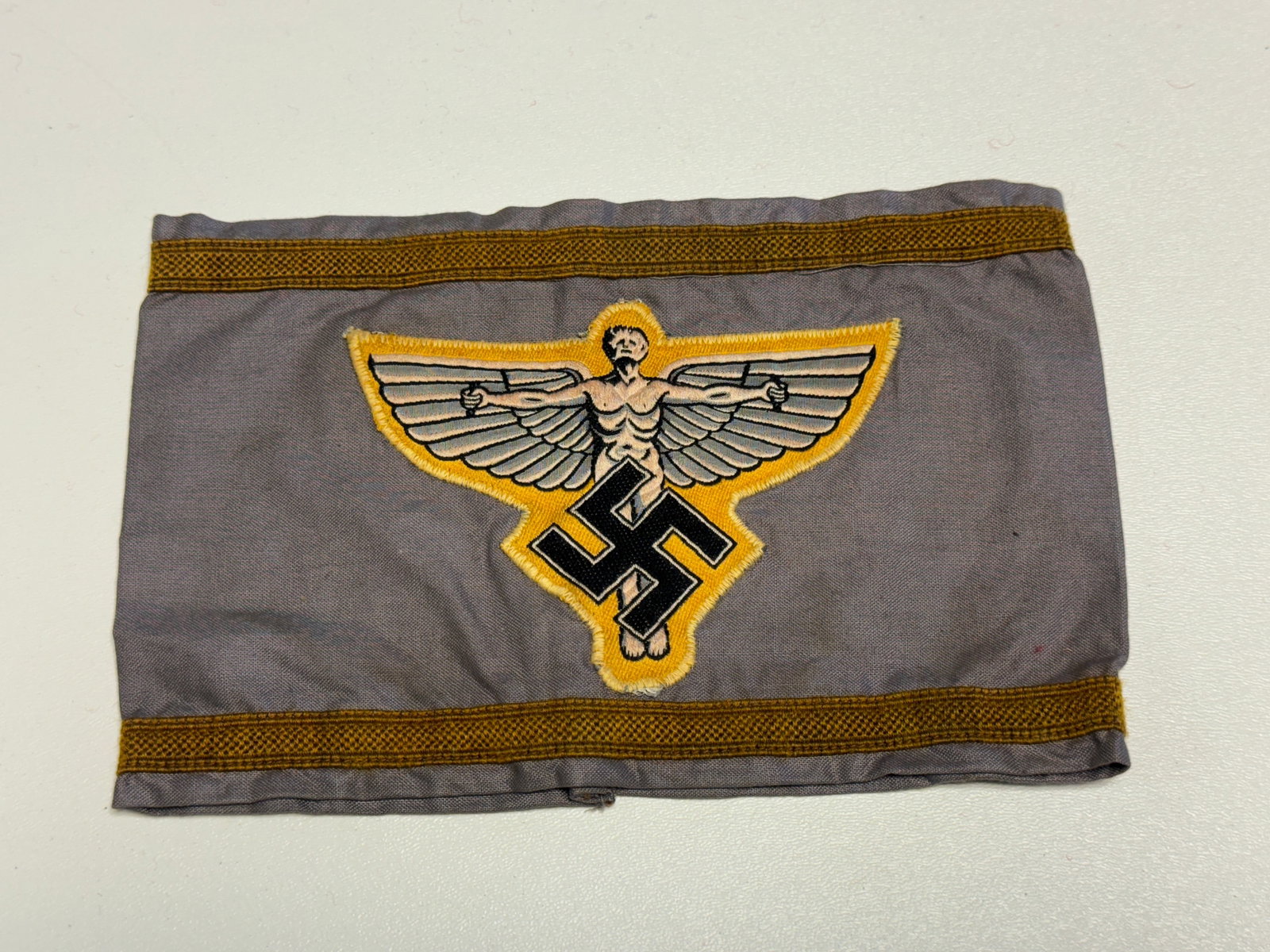 WWII German Political Organization Winged Figure Swastika Armband Insignia: WWII German political organization armband featuring a gray cloth body bordered along the upper and lower edges with decorative golden-brown woven trim. The center carries a large embroidered emblem d