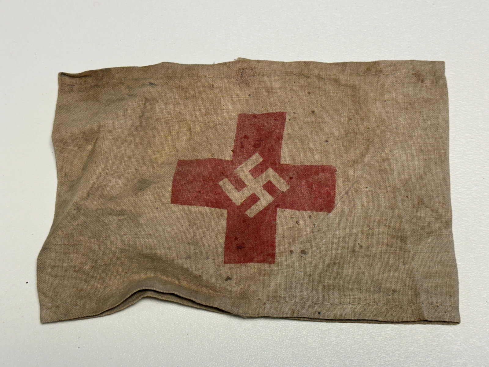 WWII German Red Cross Medical Armband with Swastika & Eagle Ink Stamp: WWII German cloth medical armband constructed from tan woven fabric and displaying a large red cross with a white swastika positioned at the center. The reverse and interior show period-style stitchin