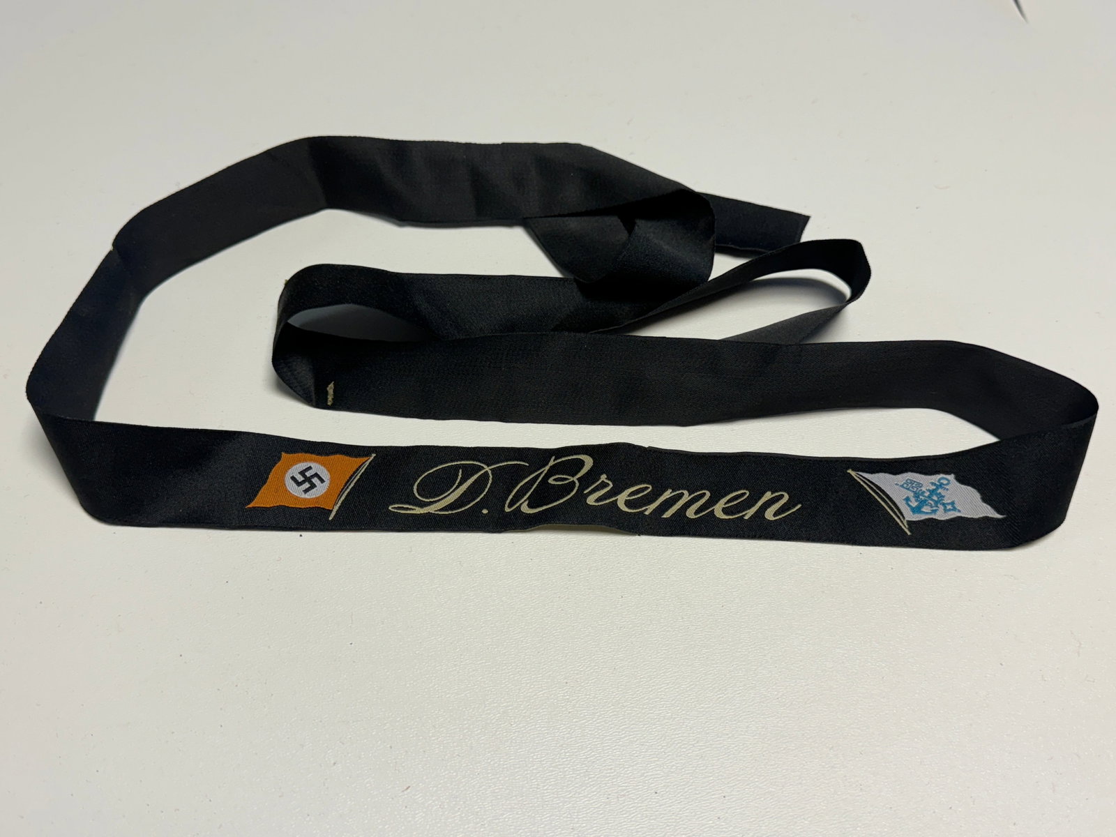WWII German Kriegsmarine “D. Bremen?? Naval Cap Tally Ribbon: WWII German naval cap tally constructed from black woven ribbon and prominently embroidered “D. Bremen” in cream-colored script. The decorated section is flanked by two embroidered flag motifs