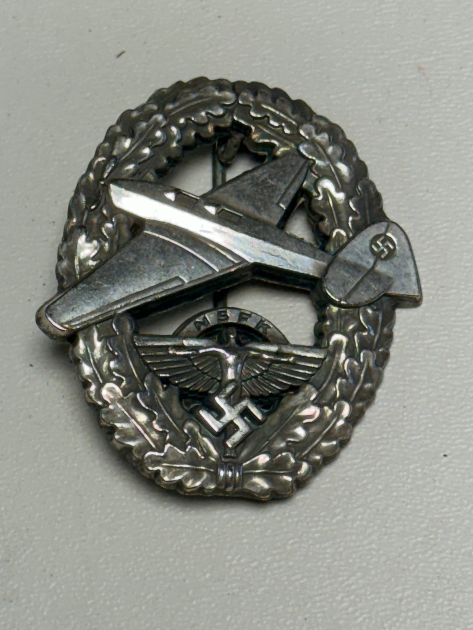 WWII German NSFK National Socialist Flying Corps Glider Pilot Badge: WWII German NSFK (Nationalsozialistisches Fliegerkorps / National Socialist Flying Corps) aviation badge featuring an oval oak-leaf wreath surrounding a prominently applied glider aircraft in flight. 