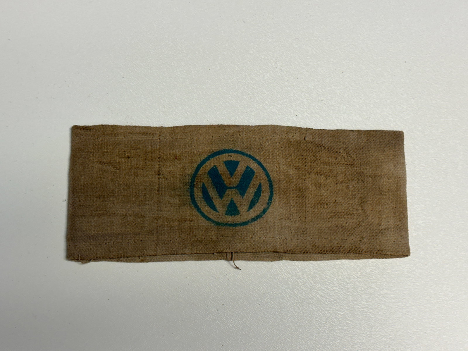WWII German Volkswagen VW Factory Worker Cloth Armband: WWII-era German Volkswagen factory worker cloth armband featuring a large blue VW emblem printed within a circular border at the center. Constructed from tan-brown woven fabric, the band displays fold
