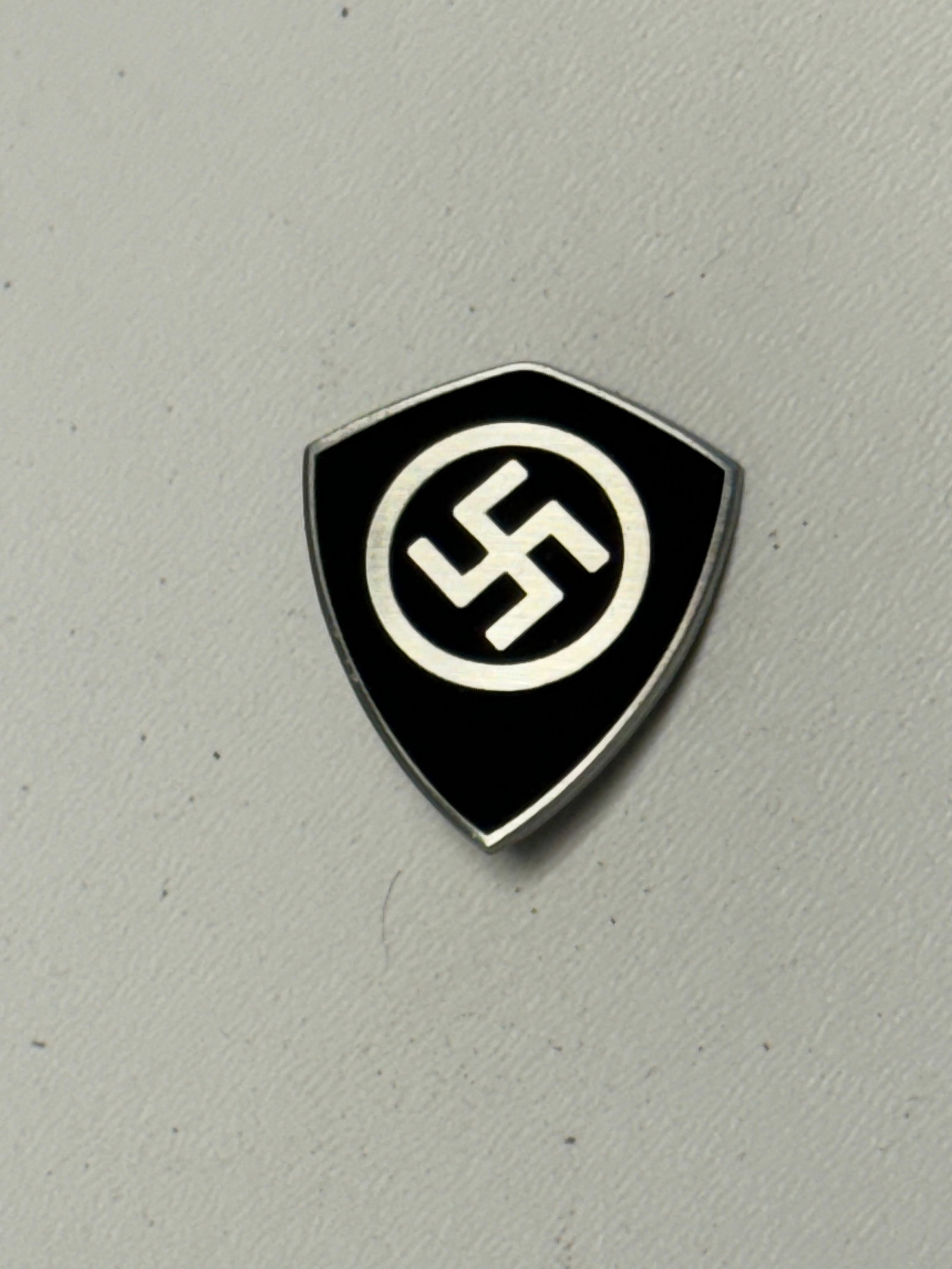 WWII German NSDAP Party Membership Shield Pin – Ges. Gesch.: WWII German NSDAP political membership shield-form pin featuring a black enamel-like field bordered in contrasting silver-colored metal, with a prominent circular emblem and swastika displayed at the 