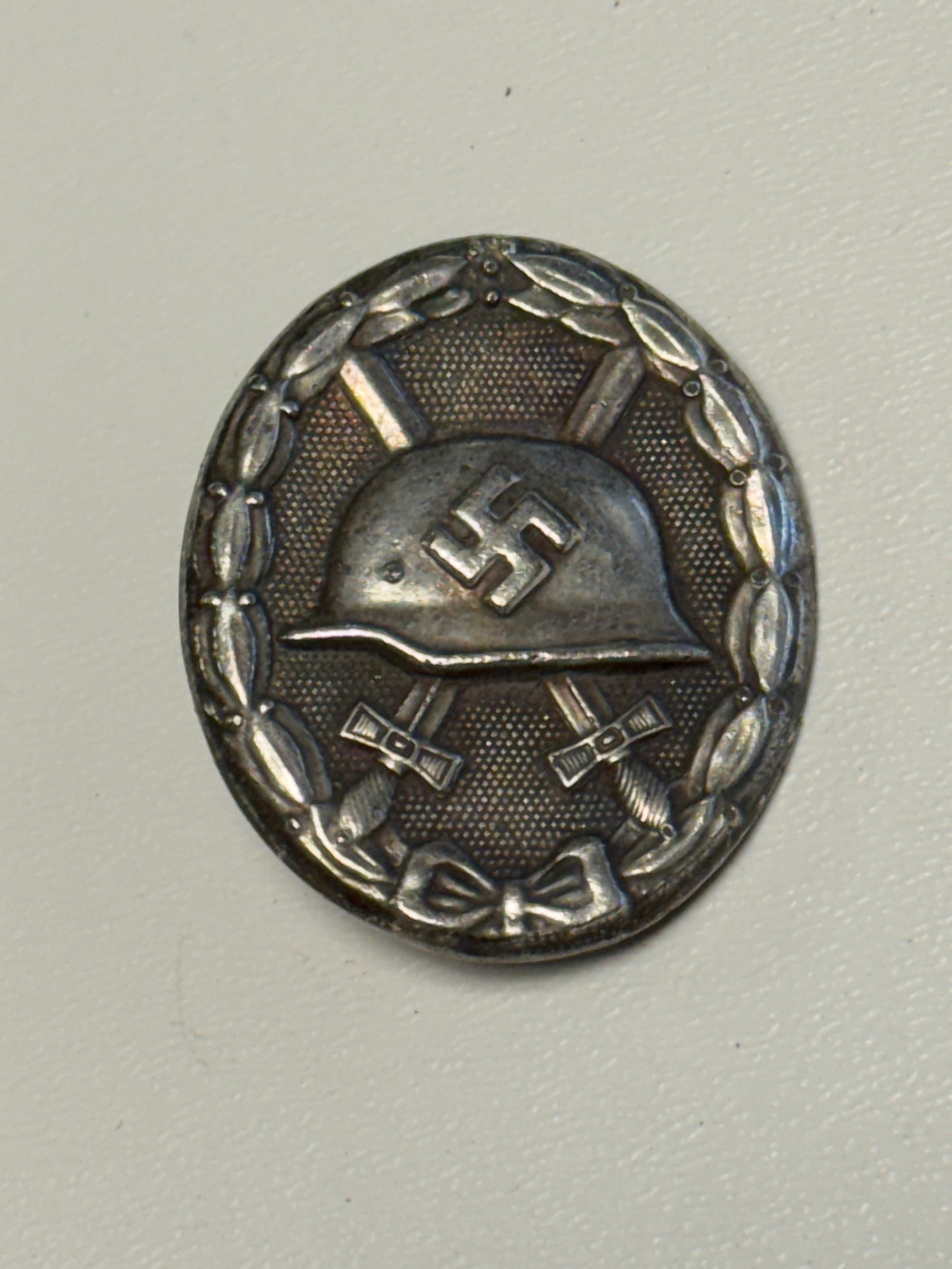 WWII German Wound Badge in Silver – Stahlhelm & Crossed Swords: WWII German Wound Badge featuring the distinctive oval form with a raised laurel wreath surrounding a central German steel helmet bearing a swastika, positioned over crossed swords against a finely te
