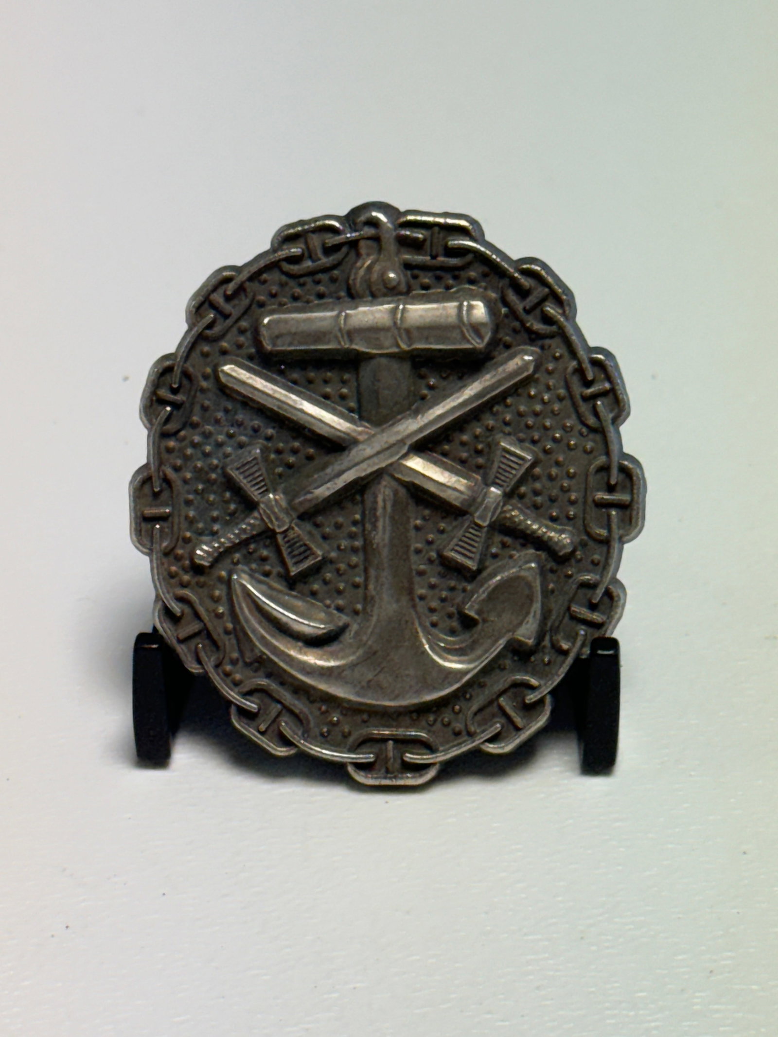 WWII German Kriegsmarine Naval Artillery Badge – Anchor & Crossed Swords: WWII German Kriegsmarine naval badge featuring a large central anchor surmounted by crossed swords, with a horizontal cylindrical element positioned above the weapons and an ornate chain-link border s