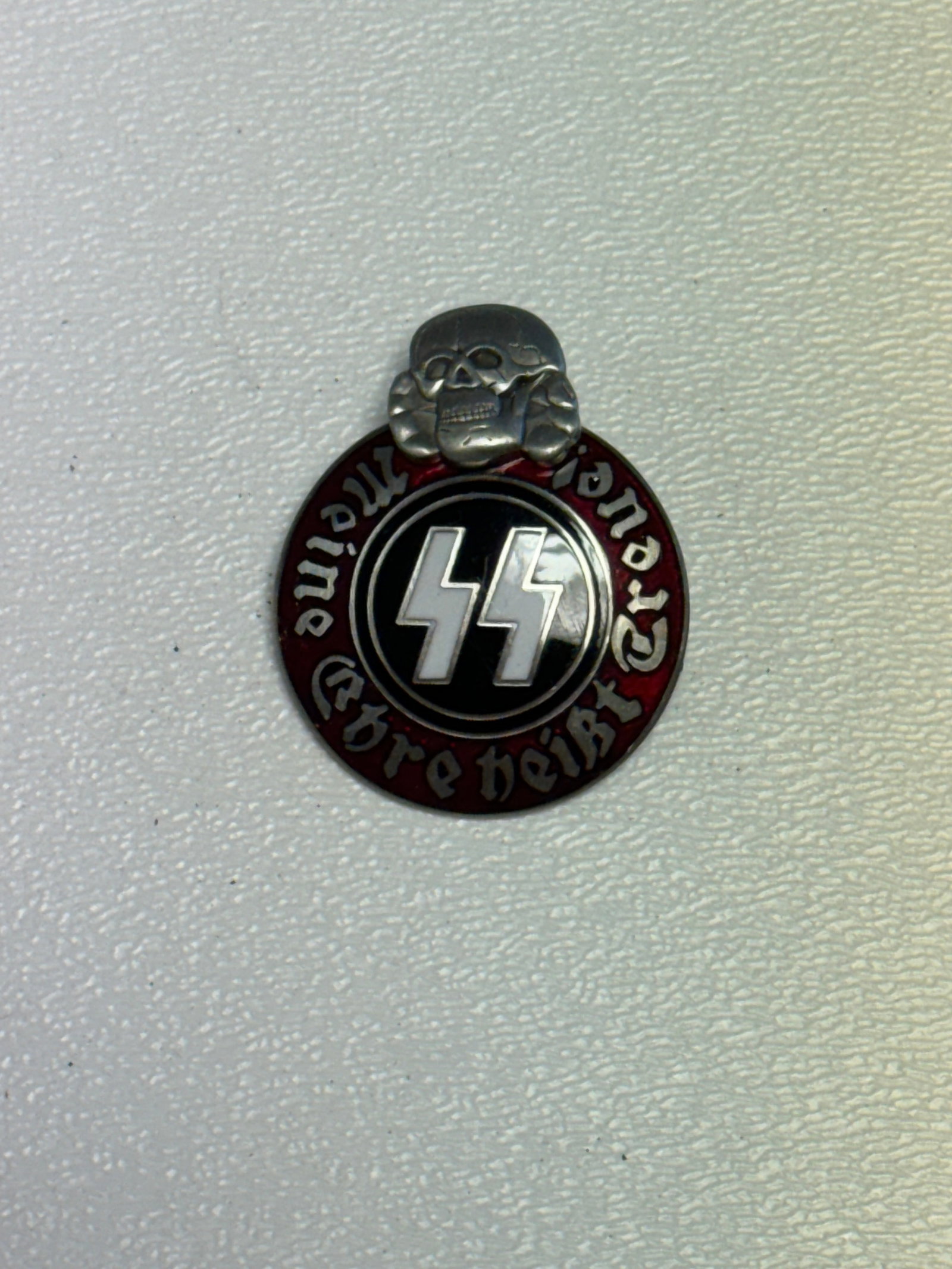 WWII German SS Totenkopf “Meine Ehre Heißt Treue?? Enamel Badge – M1/42 RZM Marked: WWII German SS-themed circular enamel badge featuring a prominent raised Totenkopf skull at the top, central black enamel field bearing the SS runes, and a deep red outer border inscribed “Meine Ehr