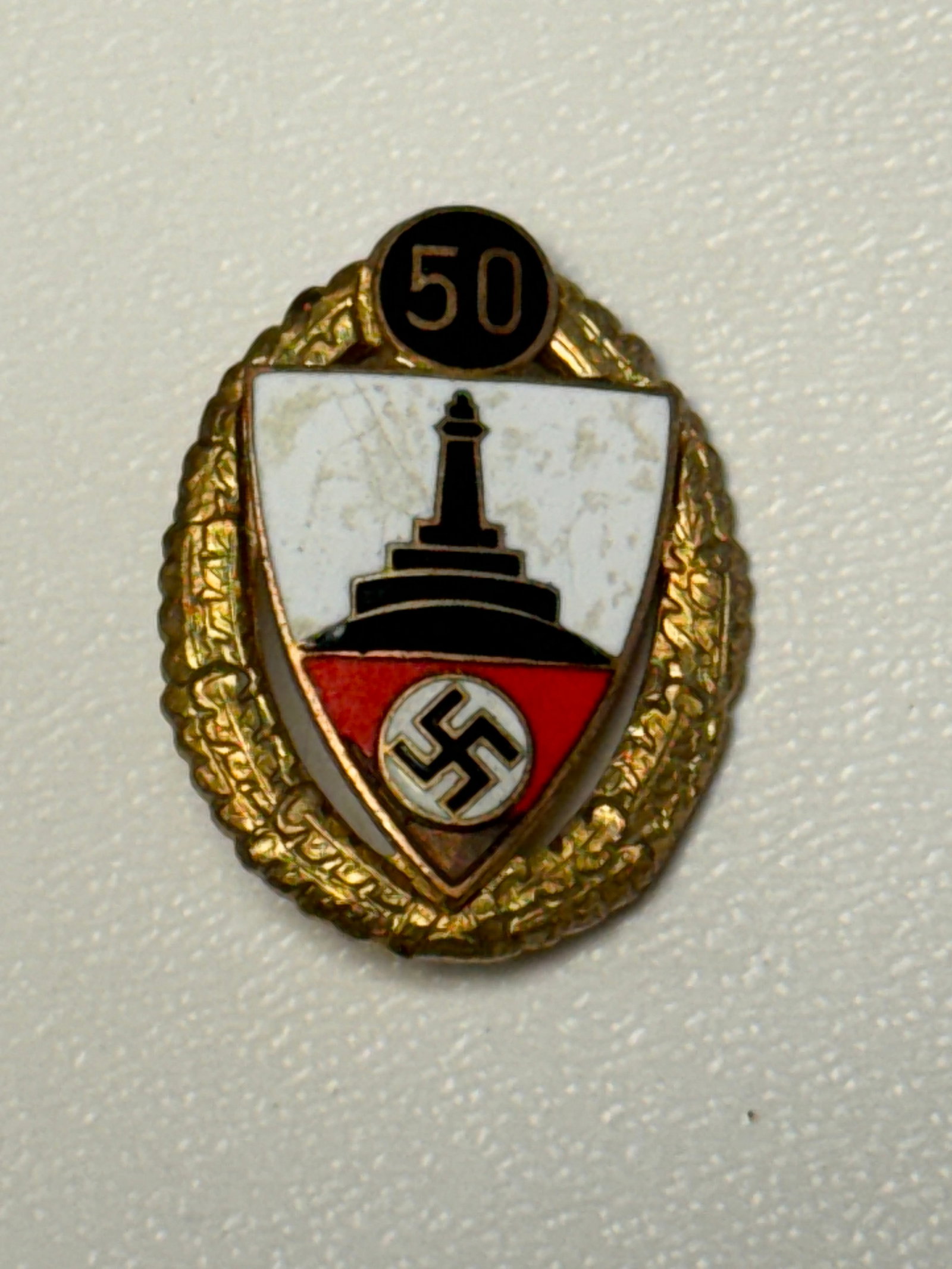 WWII German NS-RKB 50-Year Veterans Membership Honor Badge: WWII-era German veterans organization membership and service badge featuring an oval gilt wreath surrounding a white, red, and black enameled central shield. The shield displays a prominent monument m