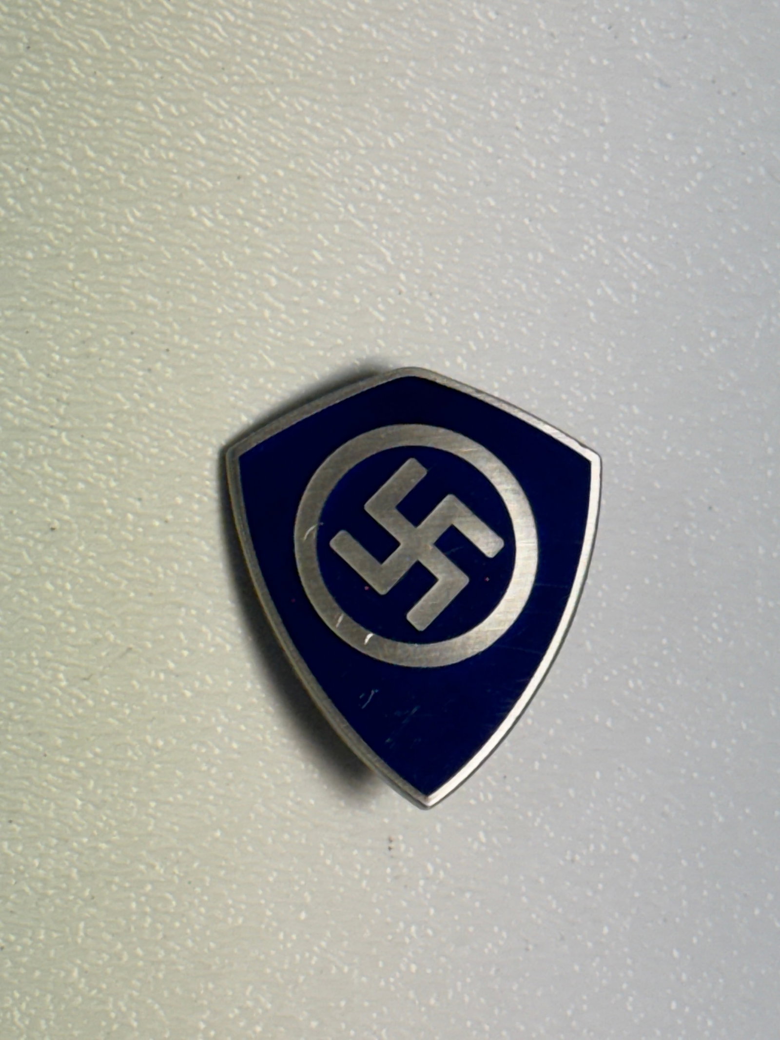 WWII German NSDAP Blue Enamel Shield Membership Badge – Ges. Gesch.: WWII-era German political organization shield-form badge featuring a deep blue enamel field bordered in silver-colored metal with a prominent central swastika enclosed within a circular ring. The comp