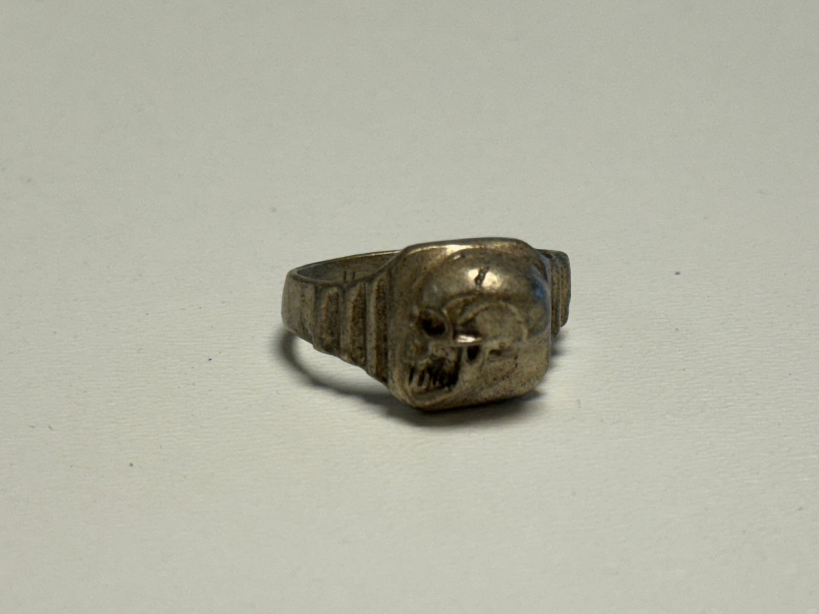 WWII German Totenkopf Skull Silver-Tone Finger Ring DRGM Marked: WWII German finger ring featuring a raised Totenkopf skull motif across the rectangular face, with deeply formed eye sockets, nasal opening, teeth, and sculpted cranial details. The shoulders are deco