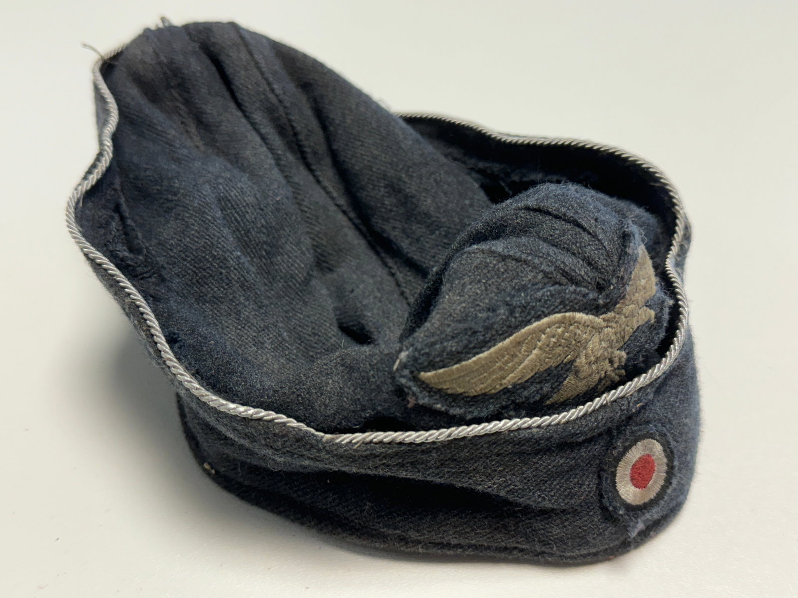 WWII German Luftwaffe Officer Overseas Side Cap with Eagle & Cockade: WWII German Luftwaffe officer’s overseas side cap constructed of dark blue-gray wool with distinctive silver-colored twisted piping running along the upper edge. The front displays an embroidered na