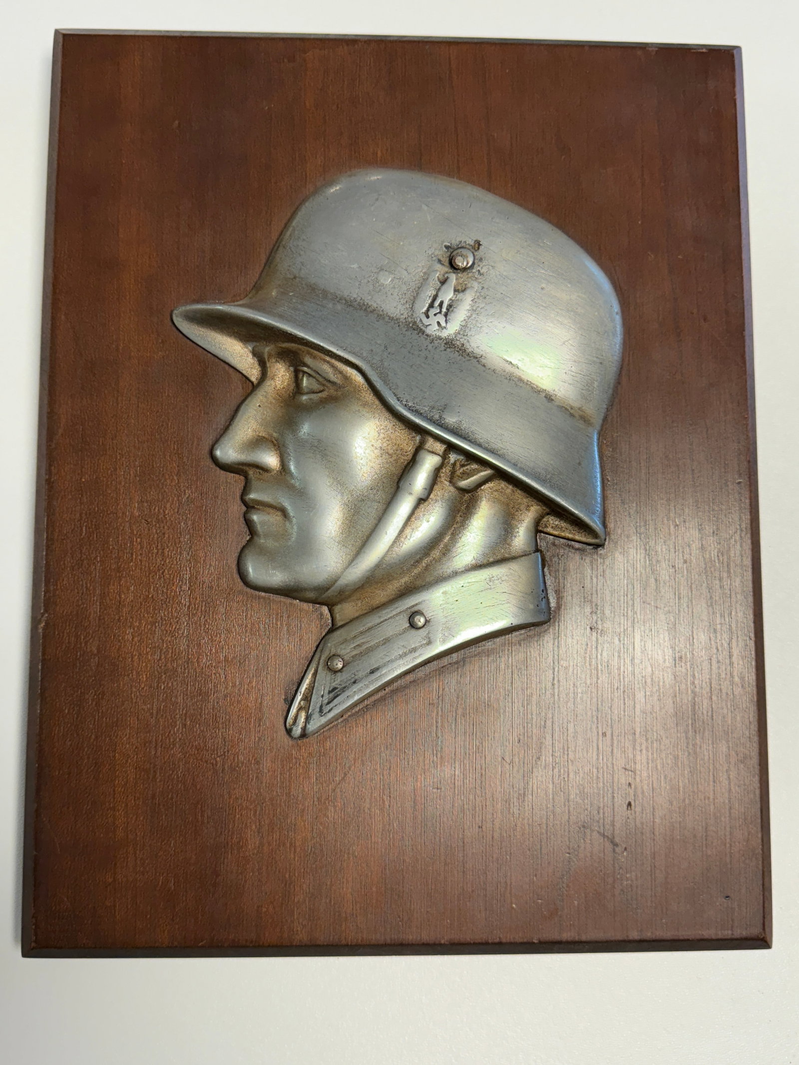 WWII German Wehrmacht Soldier Steel Helmet Profile Relief Wall Plaque: Striking WWII German military-themed wall plaque featuring a large raised metal profile portrait of a helmeted Wehrmacht soldier mounted against a dark finished wooden backing. The detailed relief dep