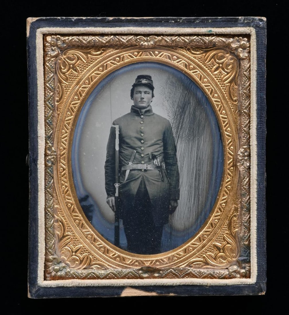 CIVIL WAR AMBROTYPE OF TRIPLE ARMED SOLDIER.: Sixth plate standing view of Union infantryman wearing 9-button frock, forage cap, holding model 1842 musket with bayonet. A Sheffield bowie knife and Colt navy style pistol are displayed in his rolle