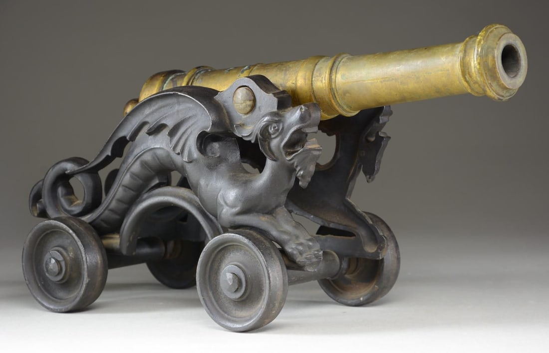 SMALL BRONZE SIGNAL CANNON WITH DECORATIVE: CAST IRON CARRIAGE. Unmarked small bronze signal cannon with 7/8" bore. Cast iron carriage featuring a decorative design of a dragon on either side, with cast iron wheels. Cannon tube measures 14 7/8"