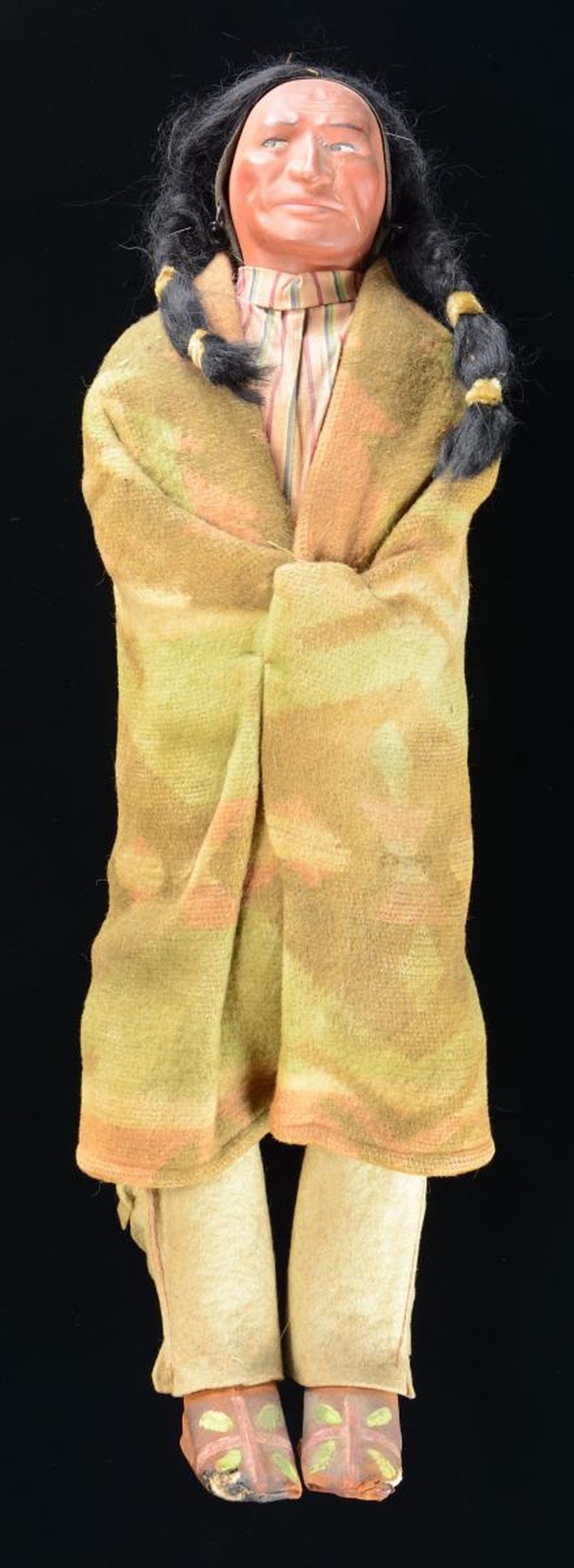 SKOOKUM NATIVE AMERICAN INDIAN DOLL.: This doll, measuring 21" in height, is an early 20th-century production figure of a Native American that was made using a composition head with hair in pigtails. The doll is wearing a striped shirt wi