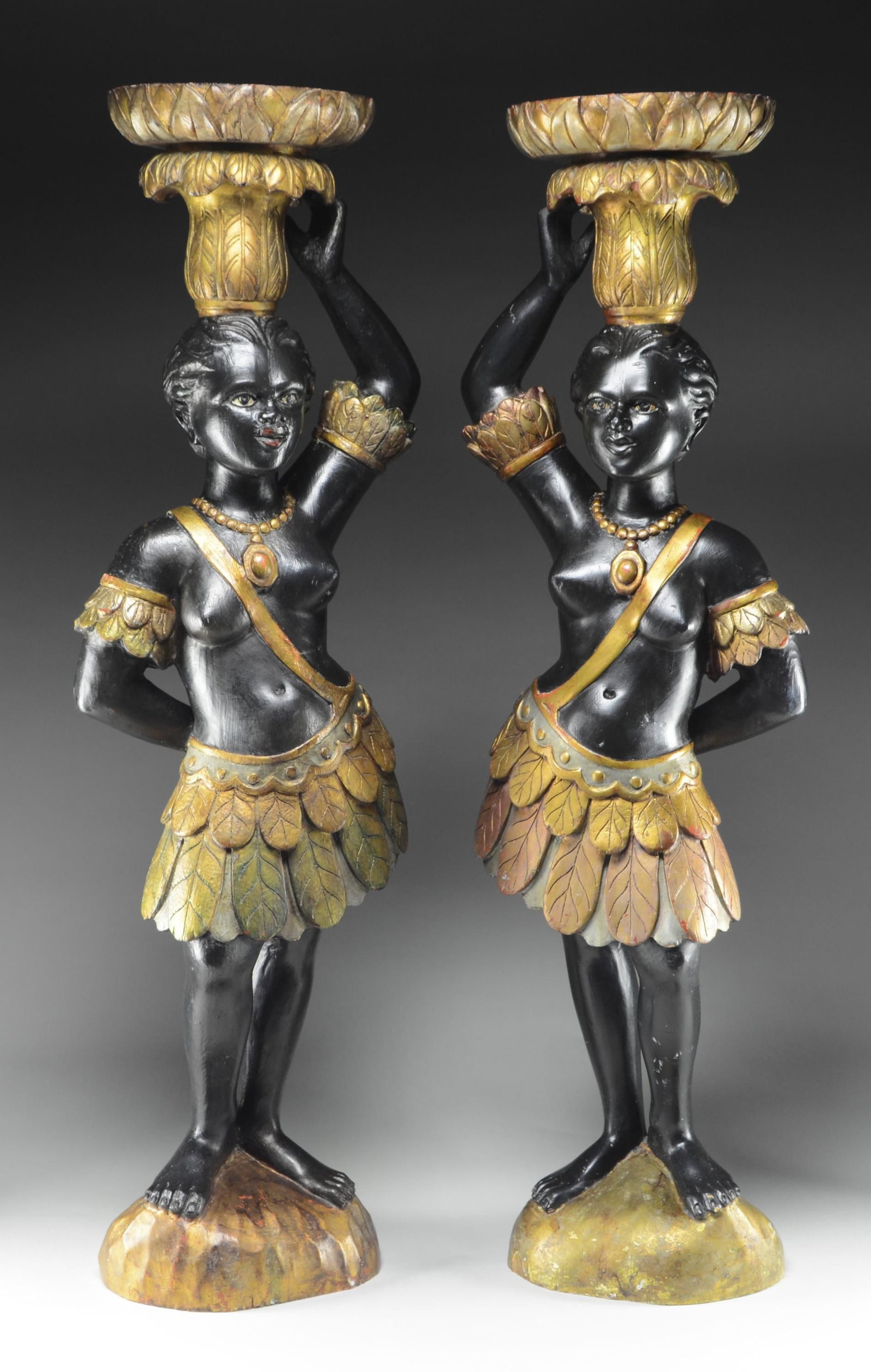 PAIR OF CARVED AND PAINTED NUBIAN STATUES.: Each figure with body in black paint, feather skirt, arm bands, and necklace in gold paint. Each with upright arm balancing a pineapple and dish. Both on round carved bases. SIZE: 52" h. CONDITION: go