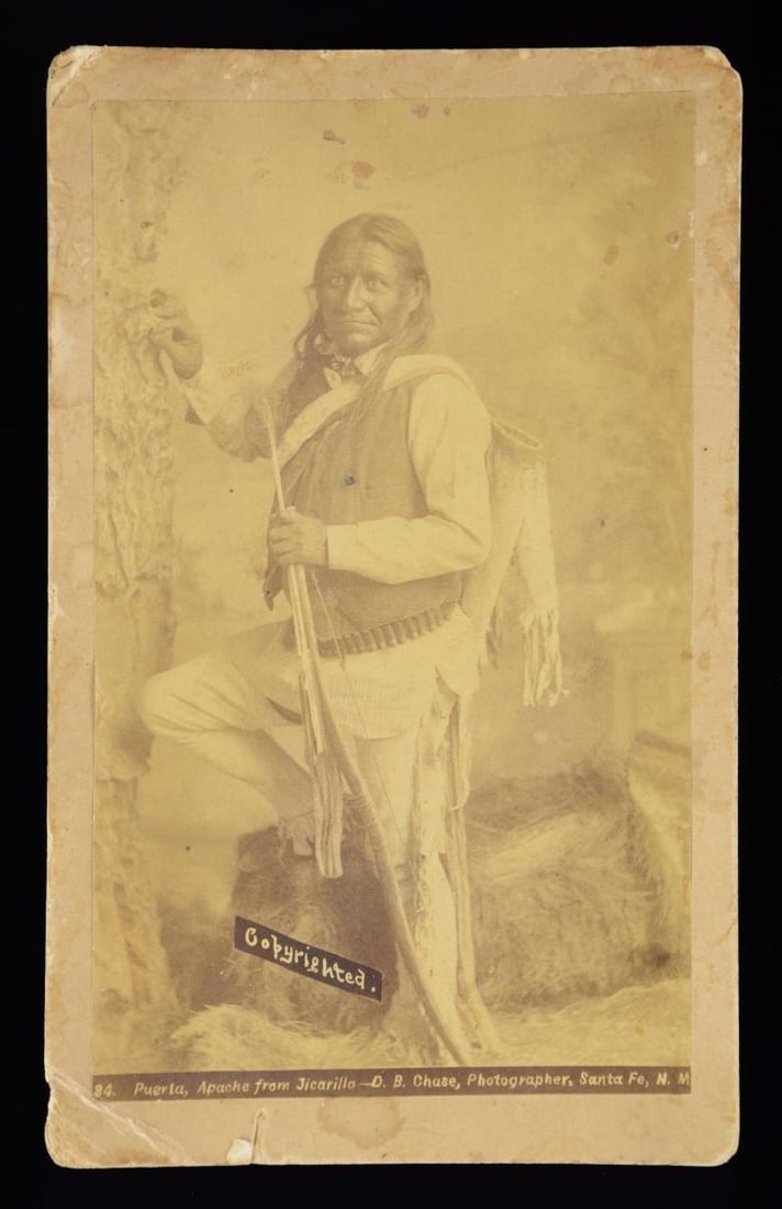 PHOTOGRAPH OF "PUERTA" JICARILLAAPACHE,: CIRCA 1890. 8" x 5" oversized image by D. B. Chase, Sante Fe, NM. Full view of Puerta standing wearing quiver and holding bow & arrows. Subject is also wearing a single loop cartridge belt and bullets
