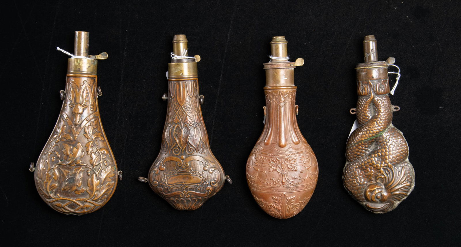 FOUR LARGE DECORATED POWDER FLASKS.: 1) 8" dolphin, Riling 1243, "BARTRAM & CO", soldered seam. 2) 8.5" acorn, oak leaves, buck's head, Riling 950, missing spring. 3) 8.5" Hawksley violin-shape, Riling 1348. 4) 8.5" "SYKES PATENT",