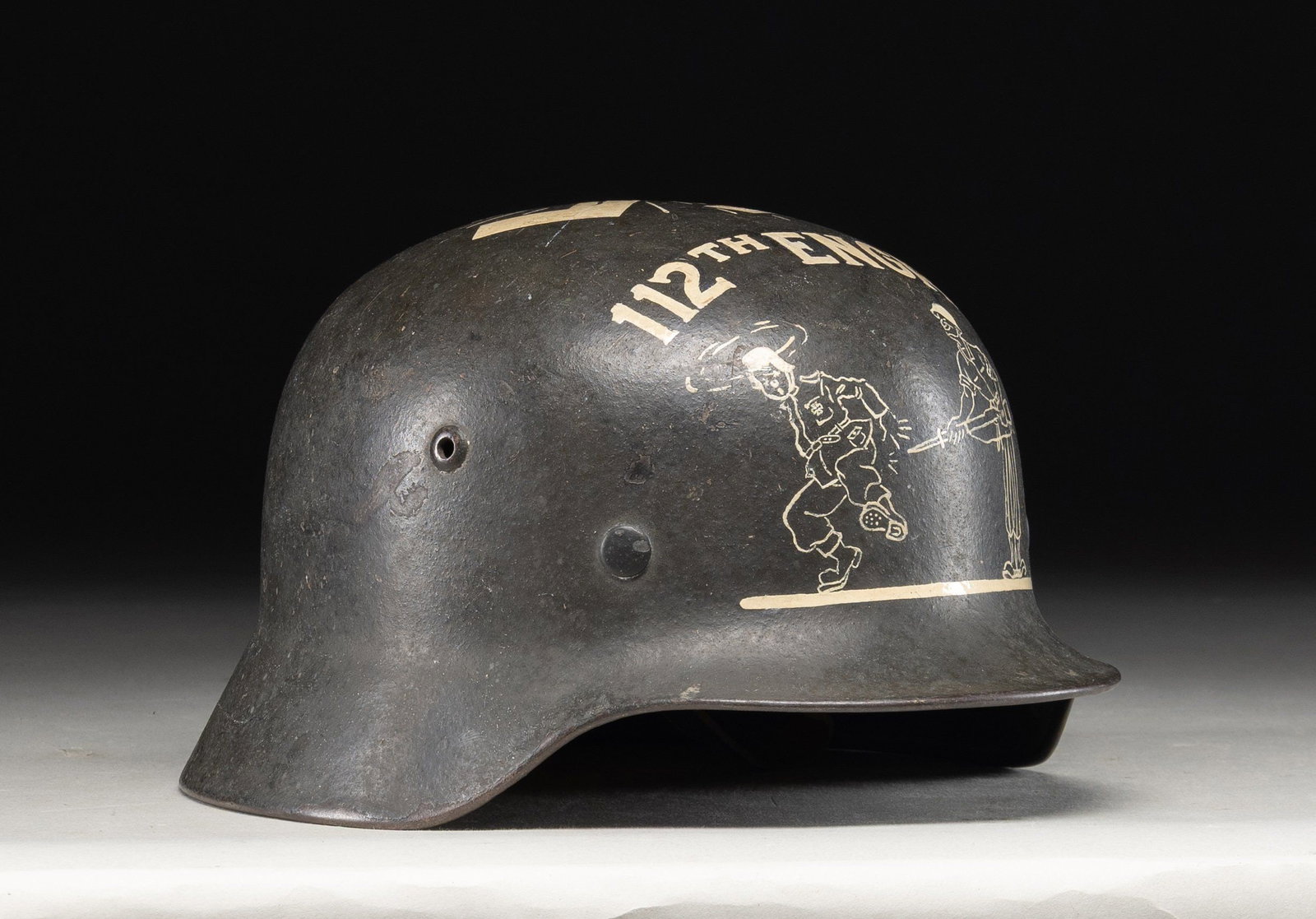 GI CAPTURED AND 112th ENGRS DECORATED GERMAN M40: STEEL HELMET. This interesting German helmet was decorated with anti German GI decorated white paint. The artwork covers the decal and depicts a well executed image of a cartoon GI poking a bayonet