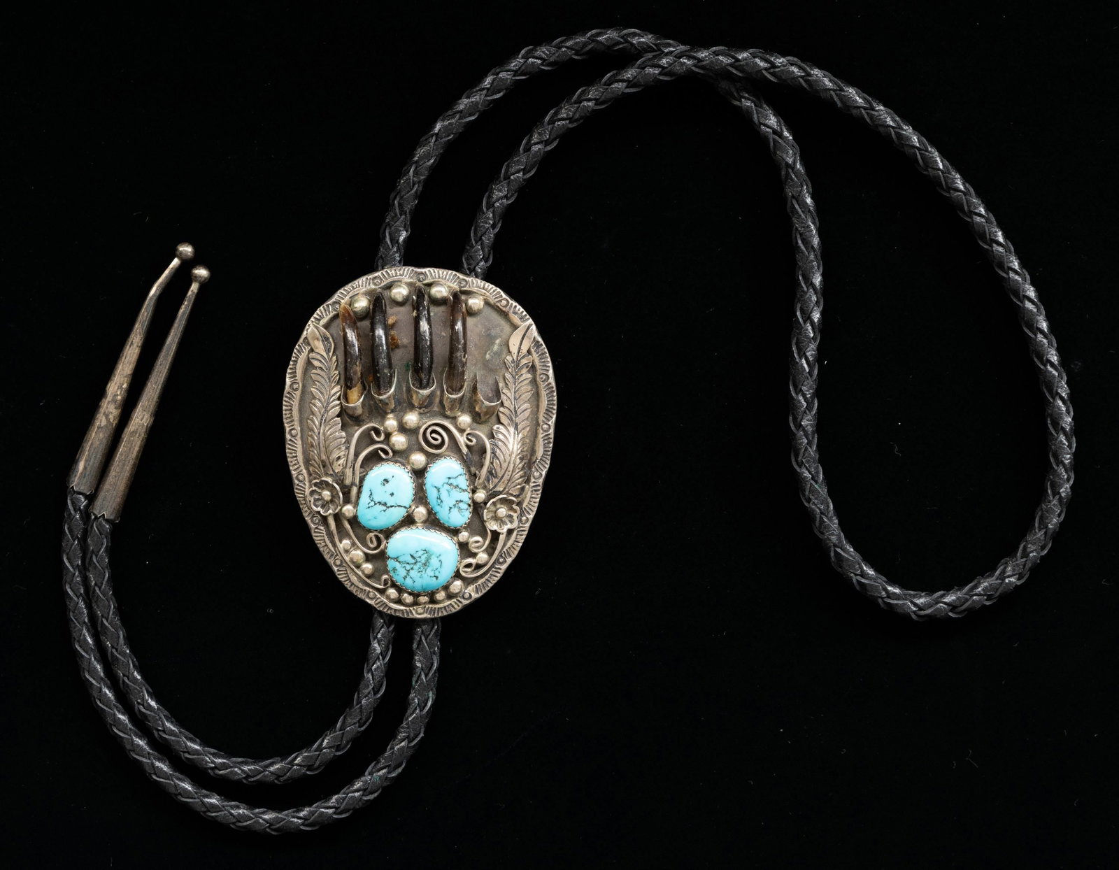 STERLING "HARRY SPENCER" NAVAJO TURQUOISE & CLAW: BOLO. 3" x 2" turquoise and small claw decoration, woven leather bolo with silver tips, signed "H. SPENCE STERLING" under curved arrow, total weight 54 grams. CONDITION: missing one of 5 small claws. 