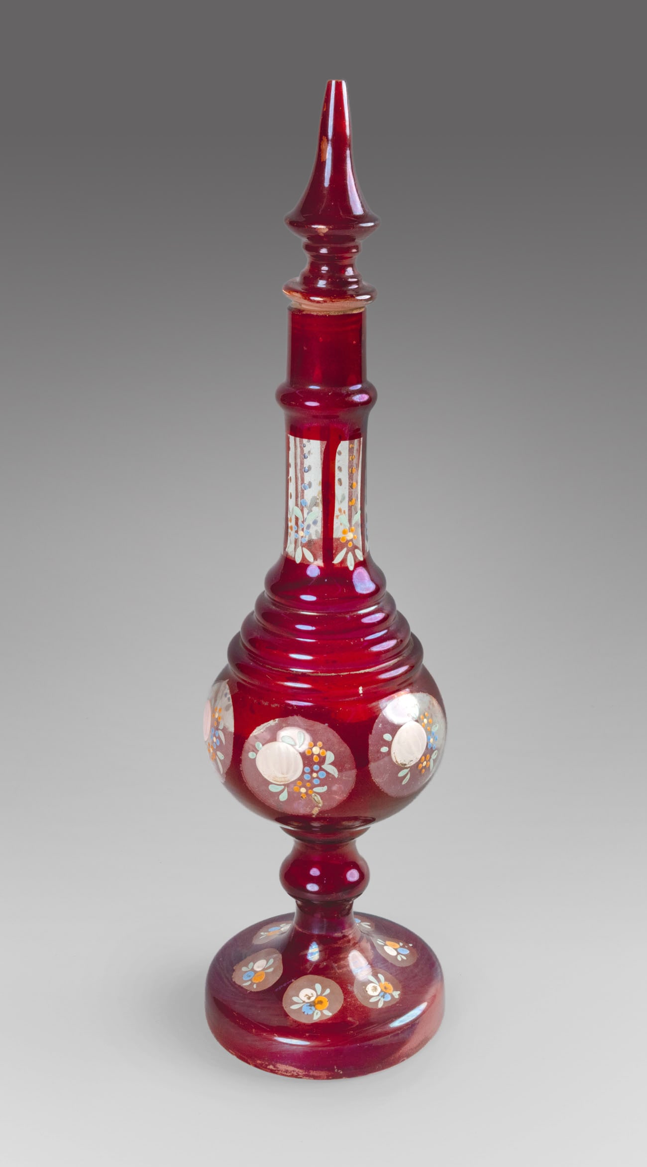 An Islamic Iridescent Ruby Glass Rosewater Sprinkler, Bohemia, 19th Century: An Islamic Iridescent Ruby Glass Rosewater Sprinkler, Bohemia, 19th Century The bulbous body set on on a thick, round base and topped by a long neck, terminating in a pointy lid. Decorated with cartou