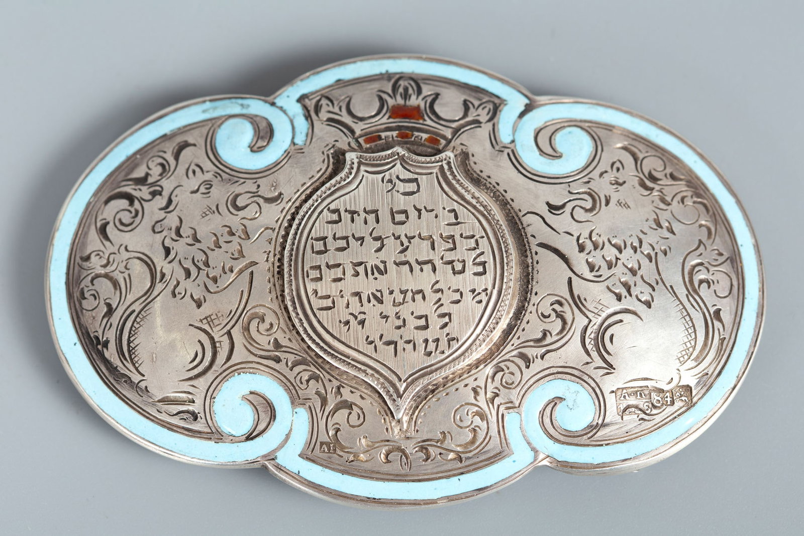 A Silver and Polychrome Enamel Yom Kippur Buckle, Russia: A Silver and Polychrome Enamel Yom Kippur Buckle, Russia Decorated with finely engraved iris motifs and a crown, highlighted with blue and red enamel. The center with an engraved relief panel depictin
