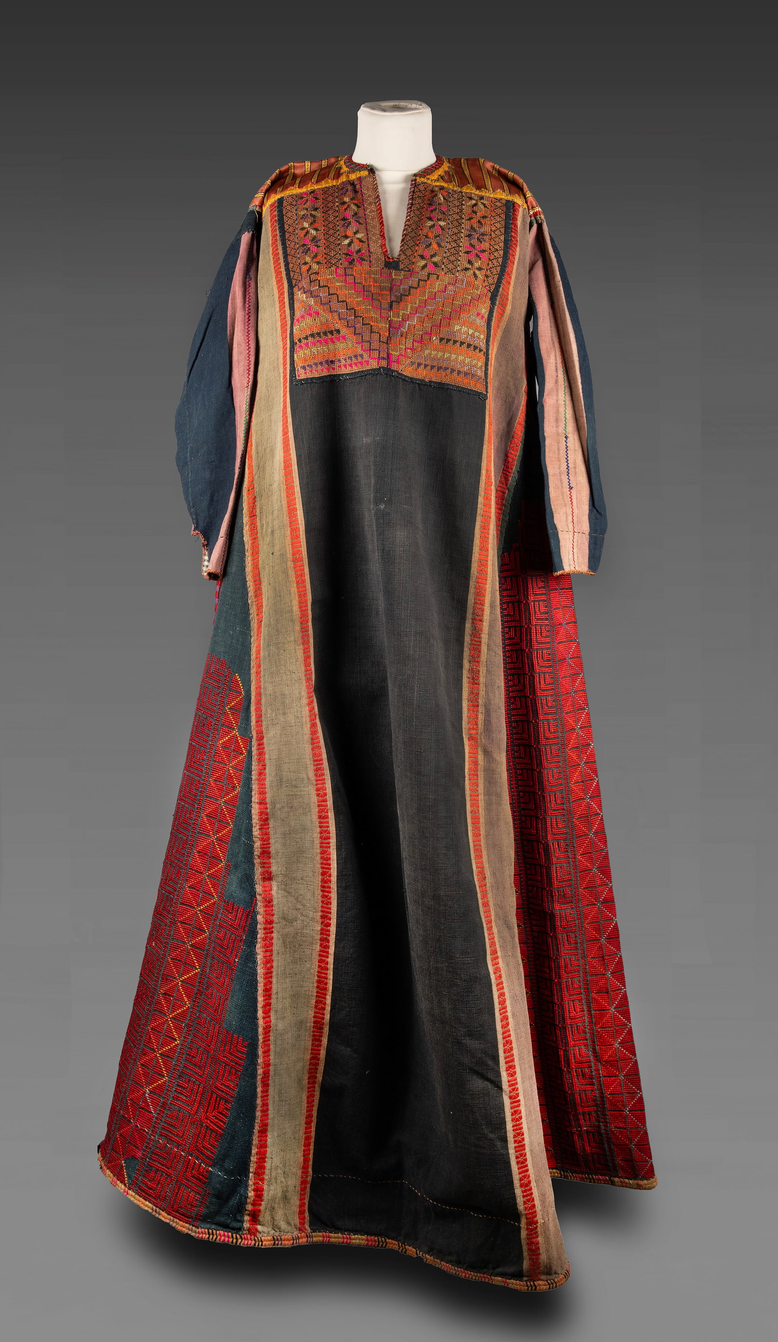 A Palestinian Embroidered Dress, Circa Early 20th Century: Composed of dark fabric with side panels in red tones featuring geometric patterns. The upper section with a densely embroidered chest panel decorated with geometric and floral motifs in multicolored 