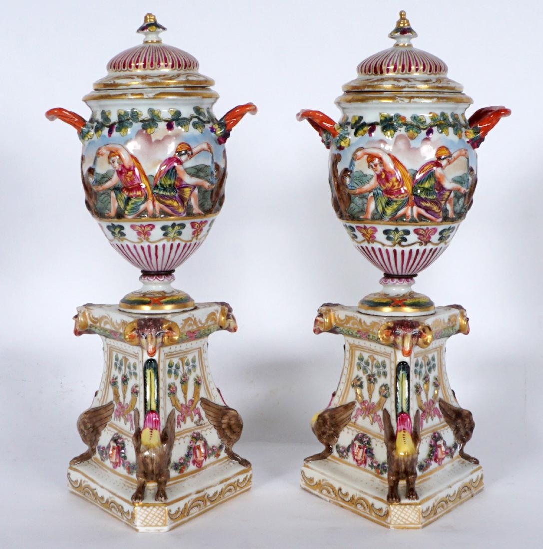 Pair Capodimonte Neoclassical Covered Urns:: PAIR CAPODIMONTE NEOCLASSICAL COVERED URNS: Floral motif finial, double handle body with relief neoclassical scene of men giving griffins. Attached to base with rams heads and griffin bodies at corner