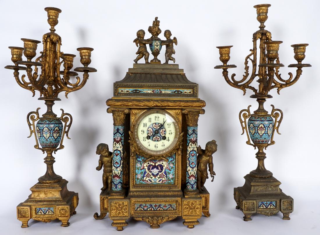 3 Piece French Champleve Clock Garniture Set: Patinated cast metal case, cherubs with urn finial, floral motif champleve enamel panels and full column supports, figural putti either side. Porcelain dial, champleve center medallion, Roman numerals