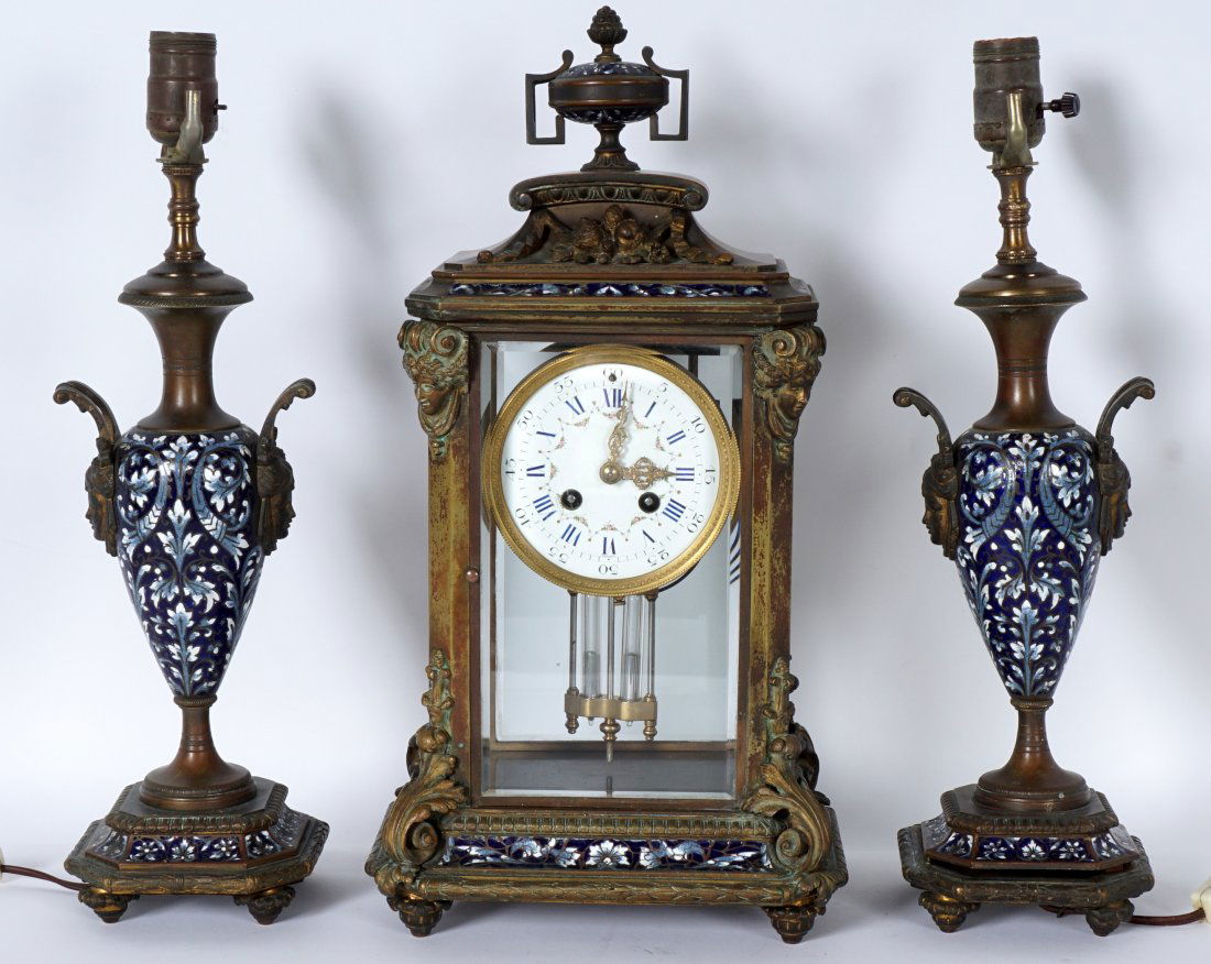 French Samuel Marti Champleve Clock Garniture Set: Three piece crystal regulator clock set. Bronze case clock applied masquerons and foliate designs, floral champleve accent panels, urn finial. Porcelain dial marked France, floral swags, Roman and Ara