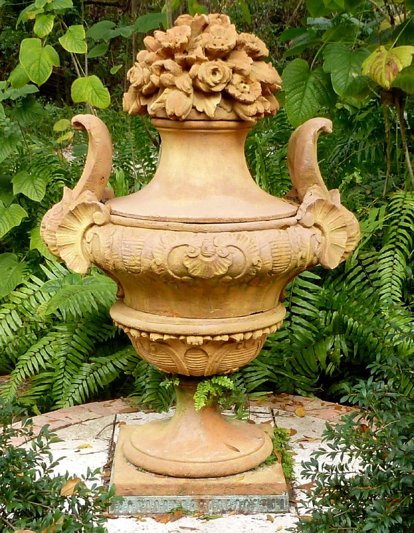 Large Terracotta Garden Urn: Removable floral bouquet top, foliate scroll handles and body, bronze panels at base. Overall 60" h. x 40.5". Condition: Signs of weathering.