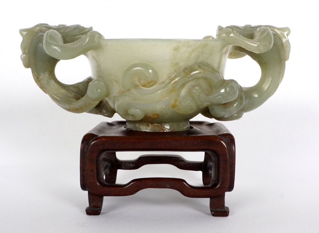 Carved Celadon Jade Cup With Dragon Handles: Diminutive libation cup with figural dragon handles, tails continuing to cup body. flared rim, cup measures 2" h. x 5". Affixed to wood stand, overall 3" h. with stand. Condition: .25" crack extending