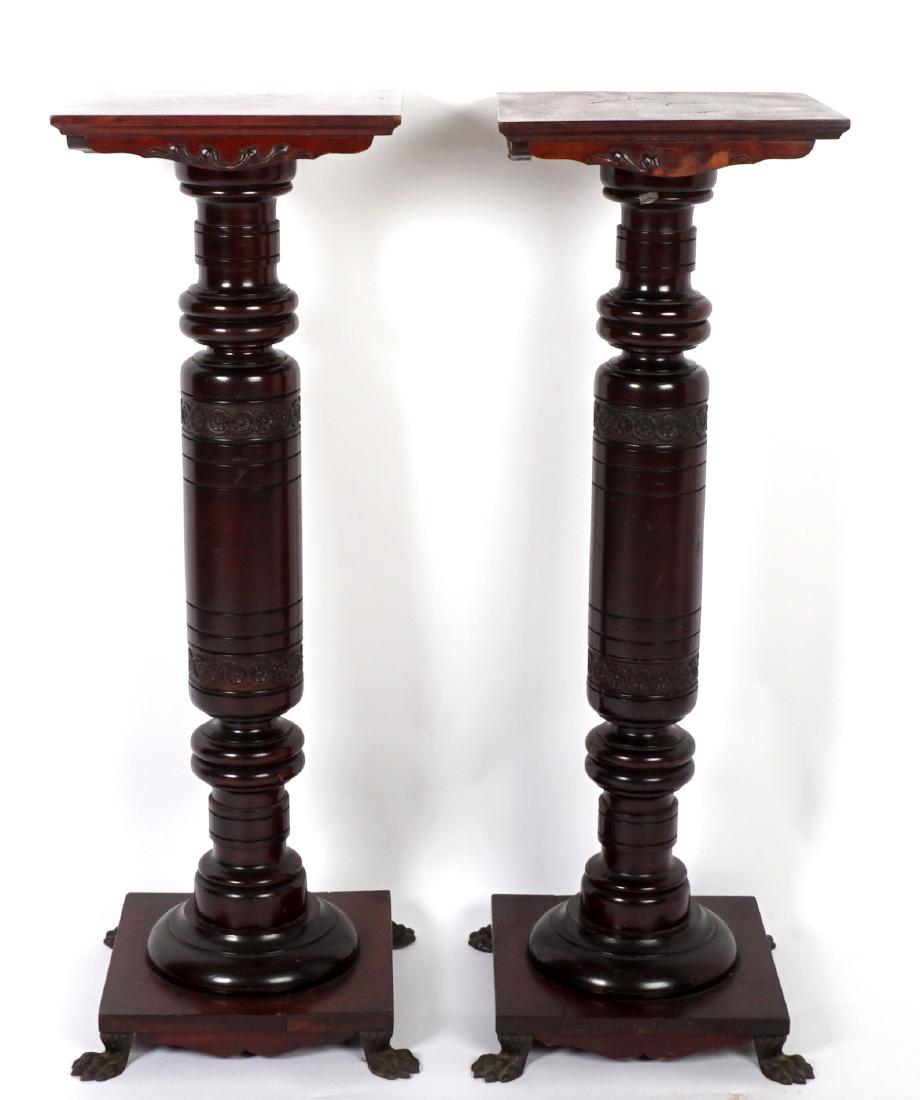 Pair Mahogany Pedestal Plant Stands: Square top over turned column pedestal support with carved floral bands, base with shaped skirt, all on four cast metal paw feet. Overall 39" h. x 13" x 13" top, 14" x 14" base. Condition: Veneer loss