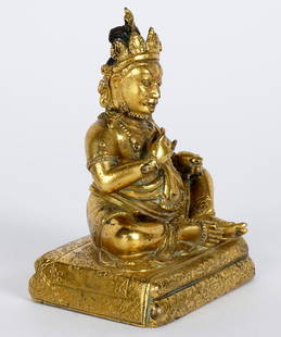 Sino Tibetan Gilt Bronze Figure of Jambhala: Heavy gilt bronze figure of seated Buddhist deity Jambhala holding mongoose in left arm, right hand in Karana Mudra position. Seated on detailed engraved pillow, figure is wearing a dhoti and adorned