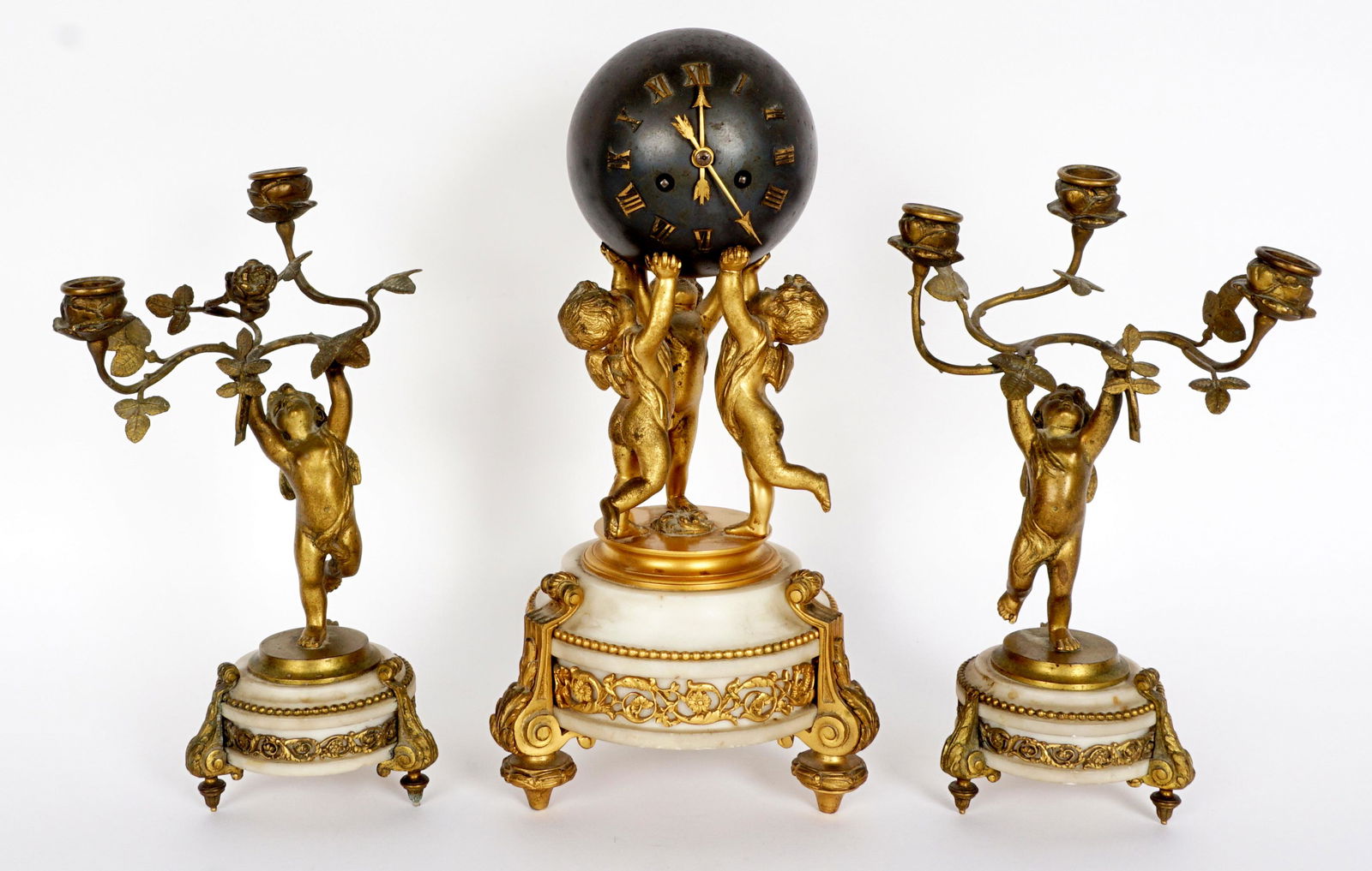 French Figural Cherub Bronze & Marble Clock: Three piece clock garniture set, metal globe clock supported by three figural gilt bronze cherubs, marble and gilt bronze base on three feet, 14.5 h. x 7". Pair matching figural bronze and marble thre