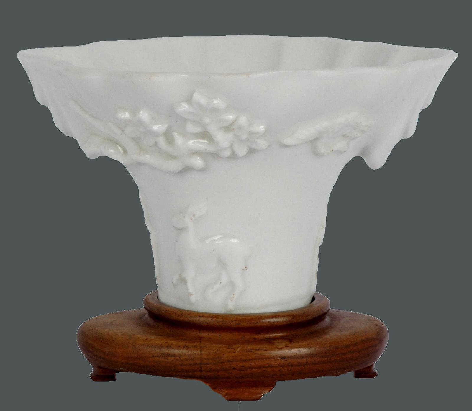 Ex-Christie's Blanc de Chine Qing Libation Cup: Qing Dynasty blanc de chine libation cup, figural deer and lion under flowering branches, scalloped rim, 3" h. x 5.25" x 5". Sold with fitted wood stand bearing a Christie's label. Condition: Rim chip
