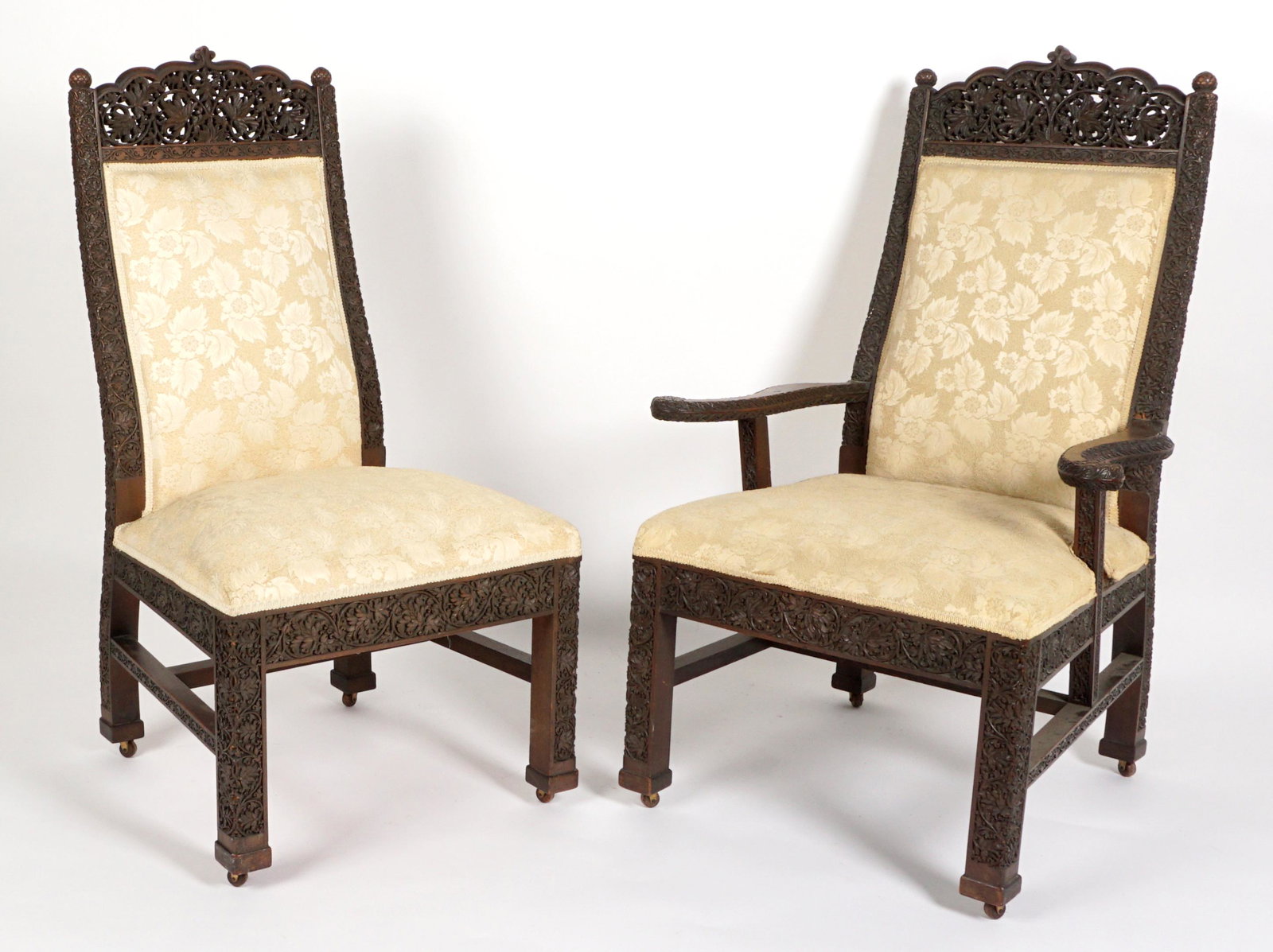 Anglo Indian Pair Pierced carved Parlor Chairs: Intricate pierced carved foliate top rail, balance of frame profusely decorated with foliate designs, on castors. Cream floral pattern upholstery. One arm chair, one side chair, arm chair measures 42.