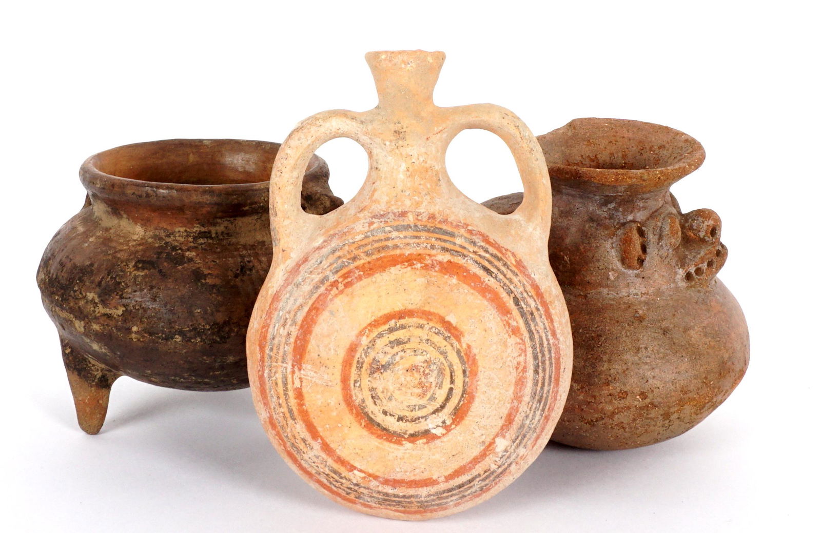 Three Pre-Columbian Pottery Vessels: An assembled collection of three pieces total to include 1) Double handled tripod vessel, traces of incised geometric designs, 4" h. x 5" dia. 2) Squat handled jug, scarified face at neck, 4.5" h. Rim