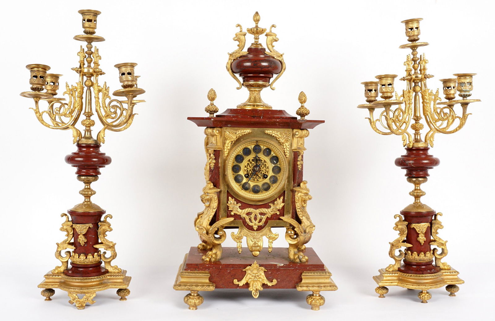 3 Piece French Marble & Gilt Bronze Mantle Clock: Three piece rouge marble and gilt bronze clock garniture set. Rouge marble clock case, urn finial with figural griffins, shaped case, gilt bronze accents, dial with blue enamel Arabic numerals, on fou