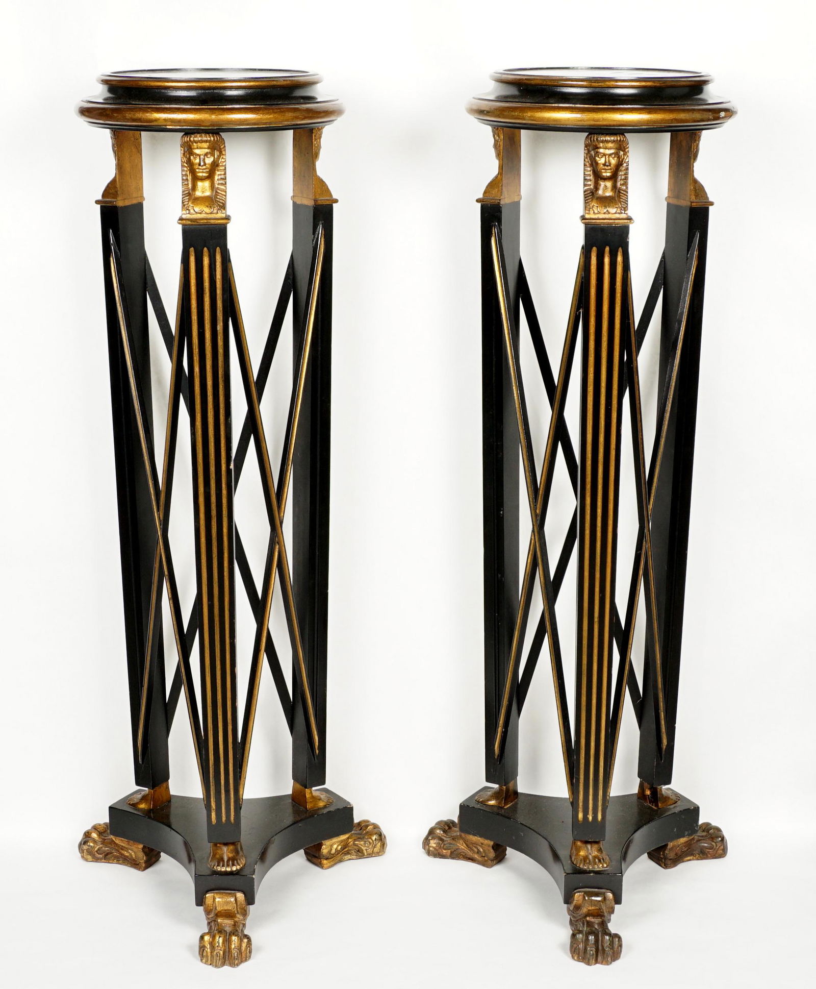 Pair Ebonized Egyptian Revival Pedestals: Ebonized and parcel gilt pedestals in the Egyptian revival style, round top with three supports, each with pharaoh busts at top, feet at base. All on splayed stretcher with paw feet. Overall 44" h. x