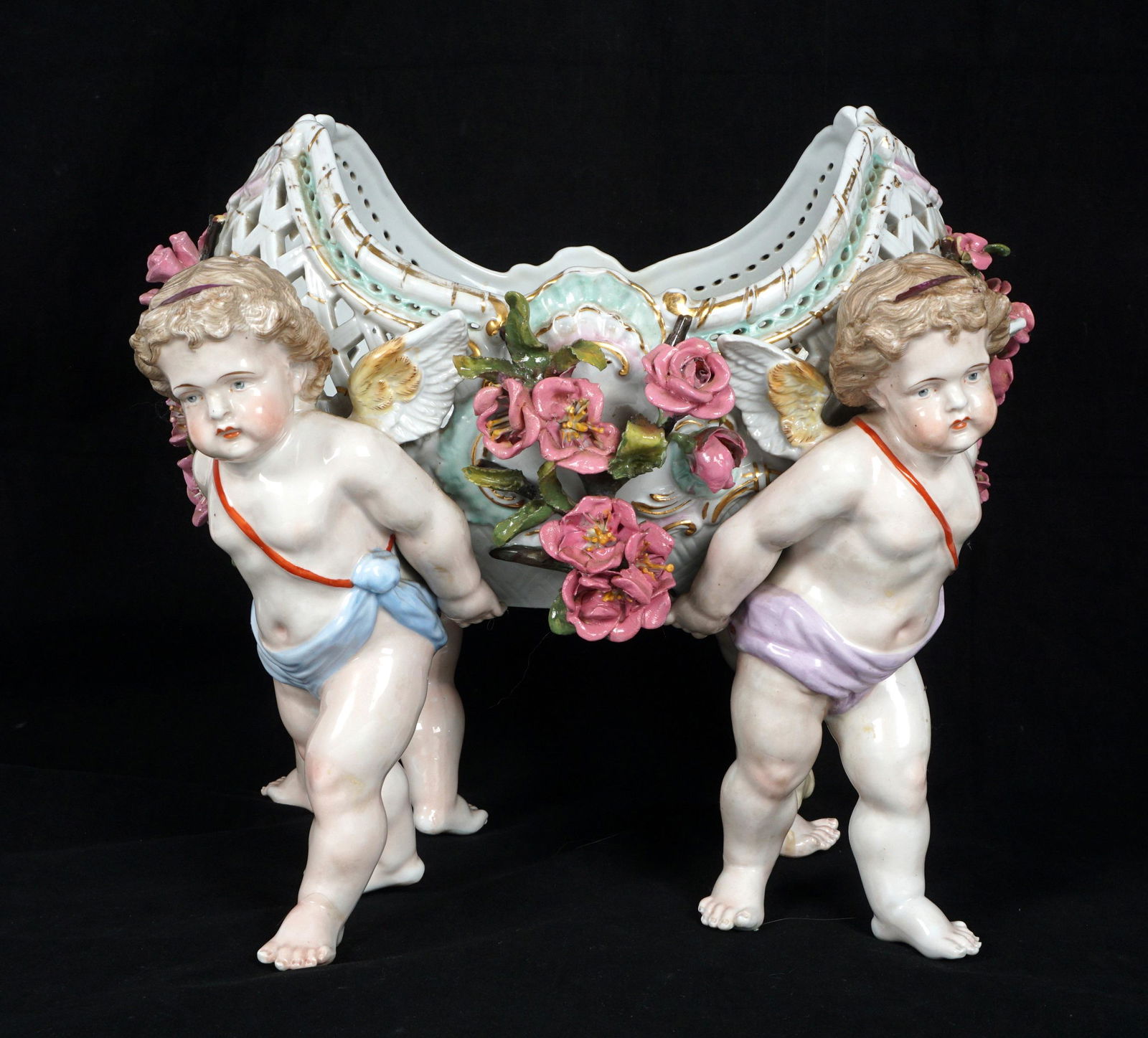 Large Schierholz Reticulated Figural Centerpiece: Four porcelain figural cherubs supporting a reticulated basket with applied flowers, marked under basket, overall 12" h. x 13" x 10.75". Condition: Extensive restoration all over, chips to flower. Sol